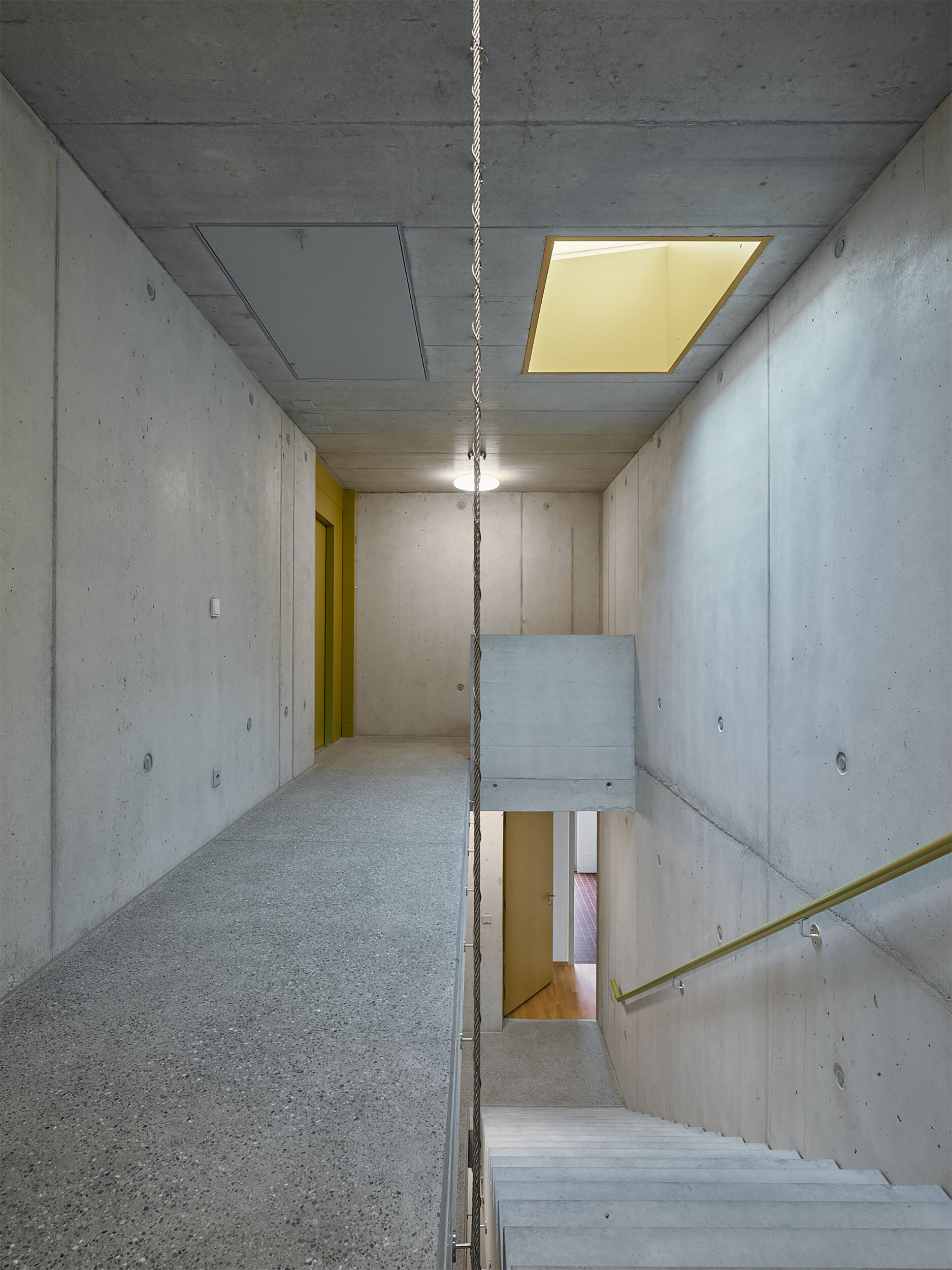 Concrete stairwell with exposed ceiling, brass handrail, and recessed skylight casting warm light