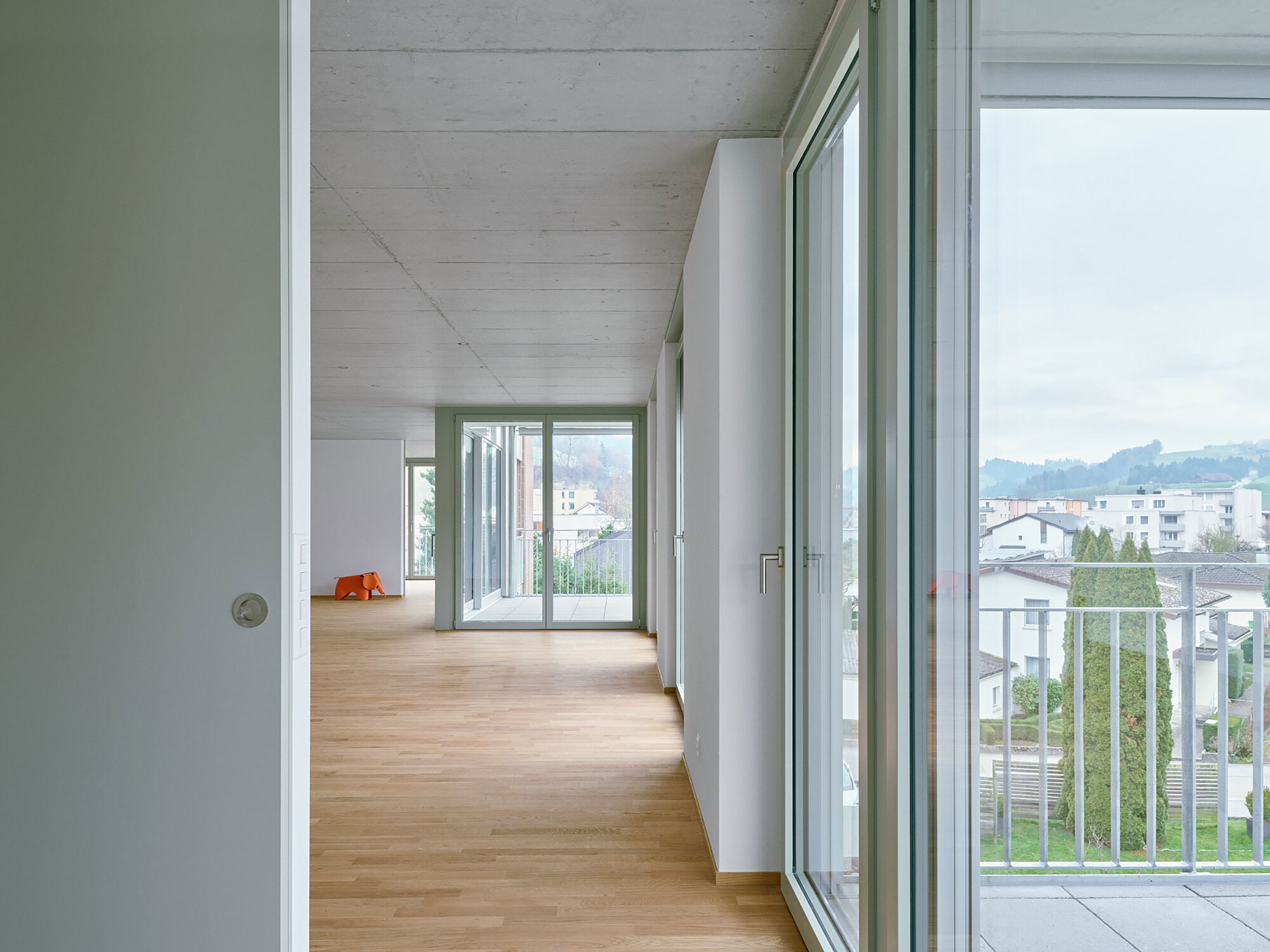 Interior corridor with light timber flooring and exposed concrete ceiling overlooking a residential neighborhood