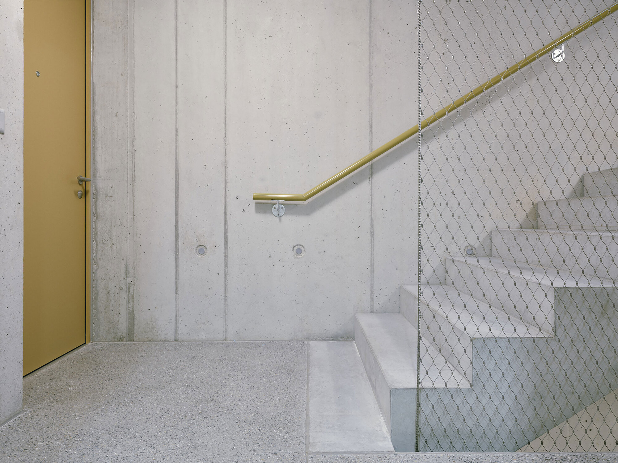 Concrete stairwell with brass handrail and wire mesh safety guard beside a mustard door