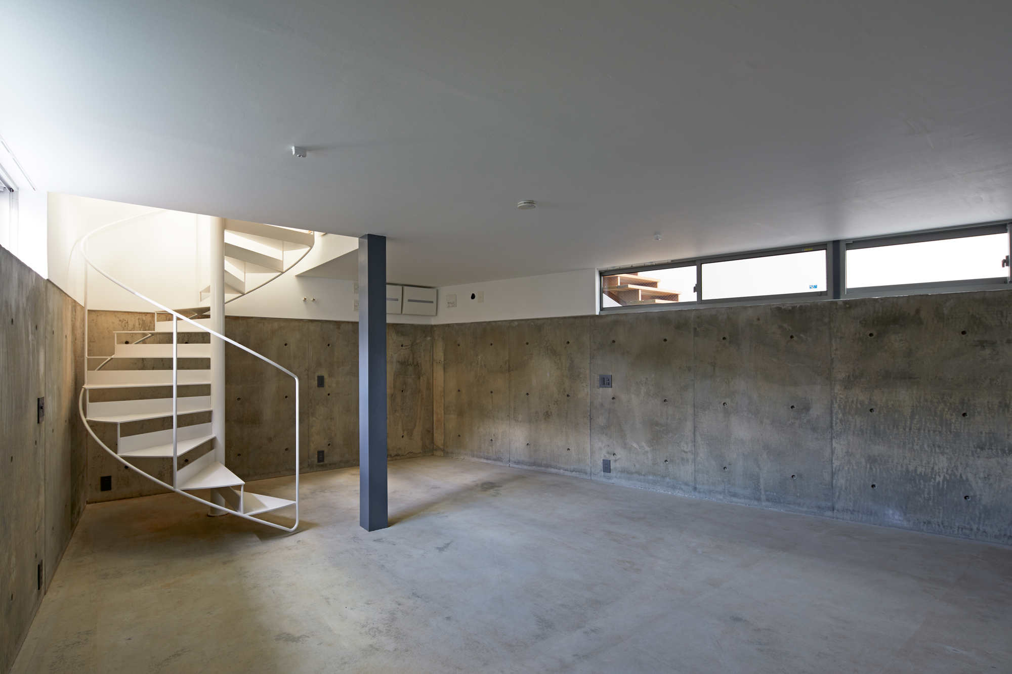 Lower level space with board-formed concrete walls, spiral stair, and steel column under clerestory windows