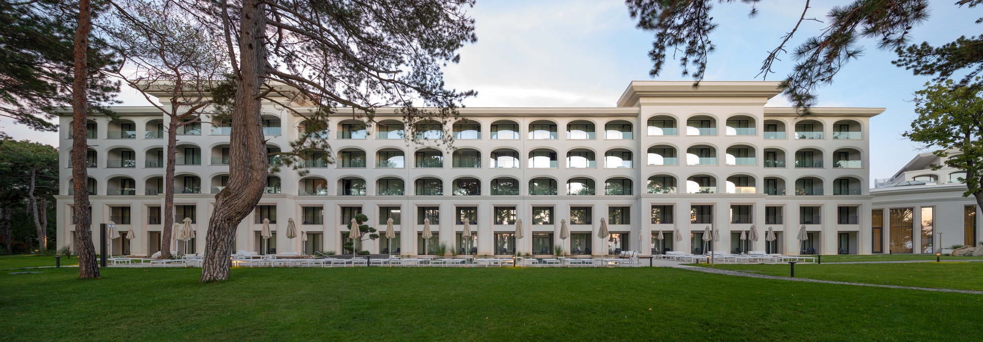 Long facade of arched balconies across the lawn with pines in the foreground