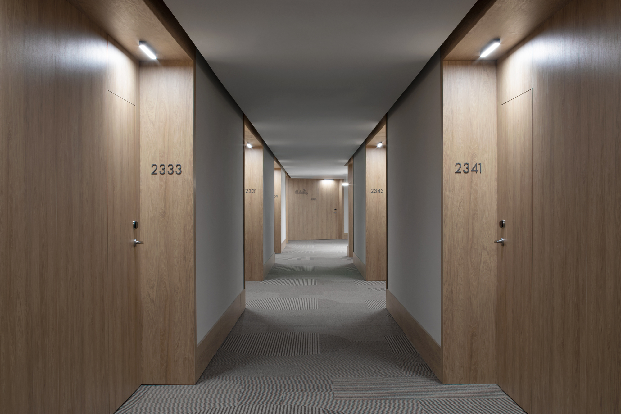 Hotel corridor with numbered wood doors and grey walls under recessed linear lighting