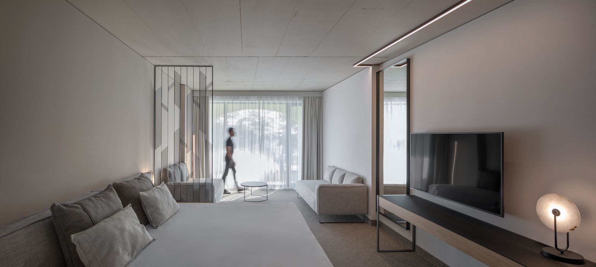 Hotel room interior with grey textiles and perforated metal partition separating bed from seating area