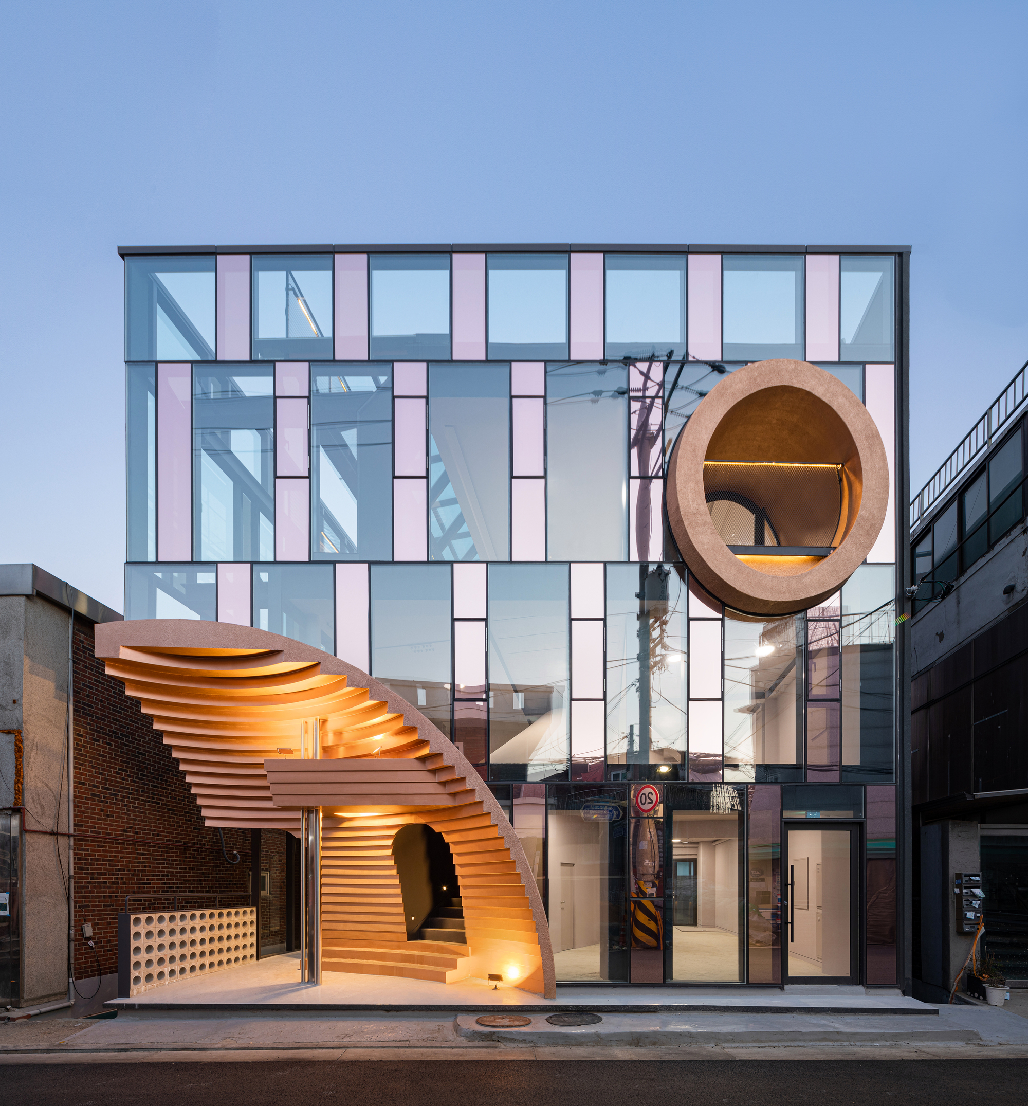 Street facade with vertical glass panels and two curved timber volumes glowing at twilight