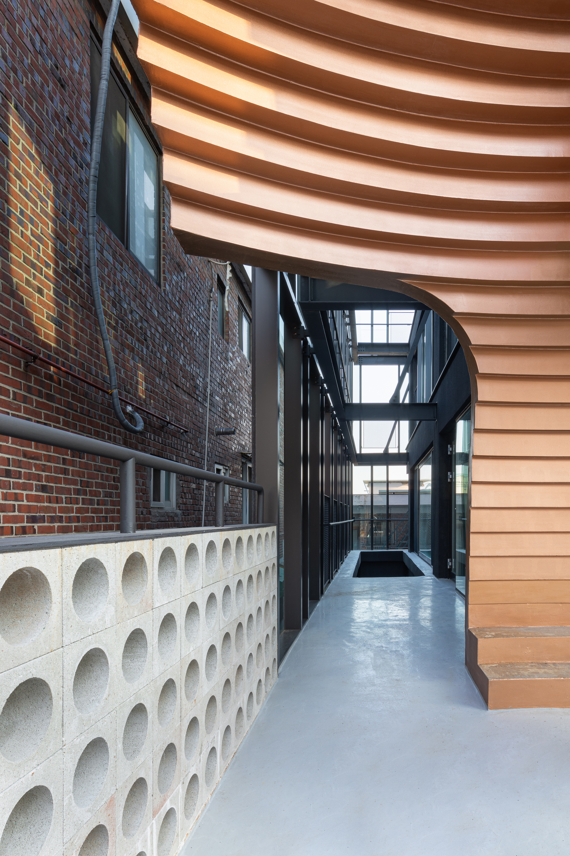 Narrow passageway between perforated concrete wall and layered timber canopy under daylight