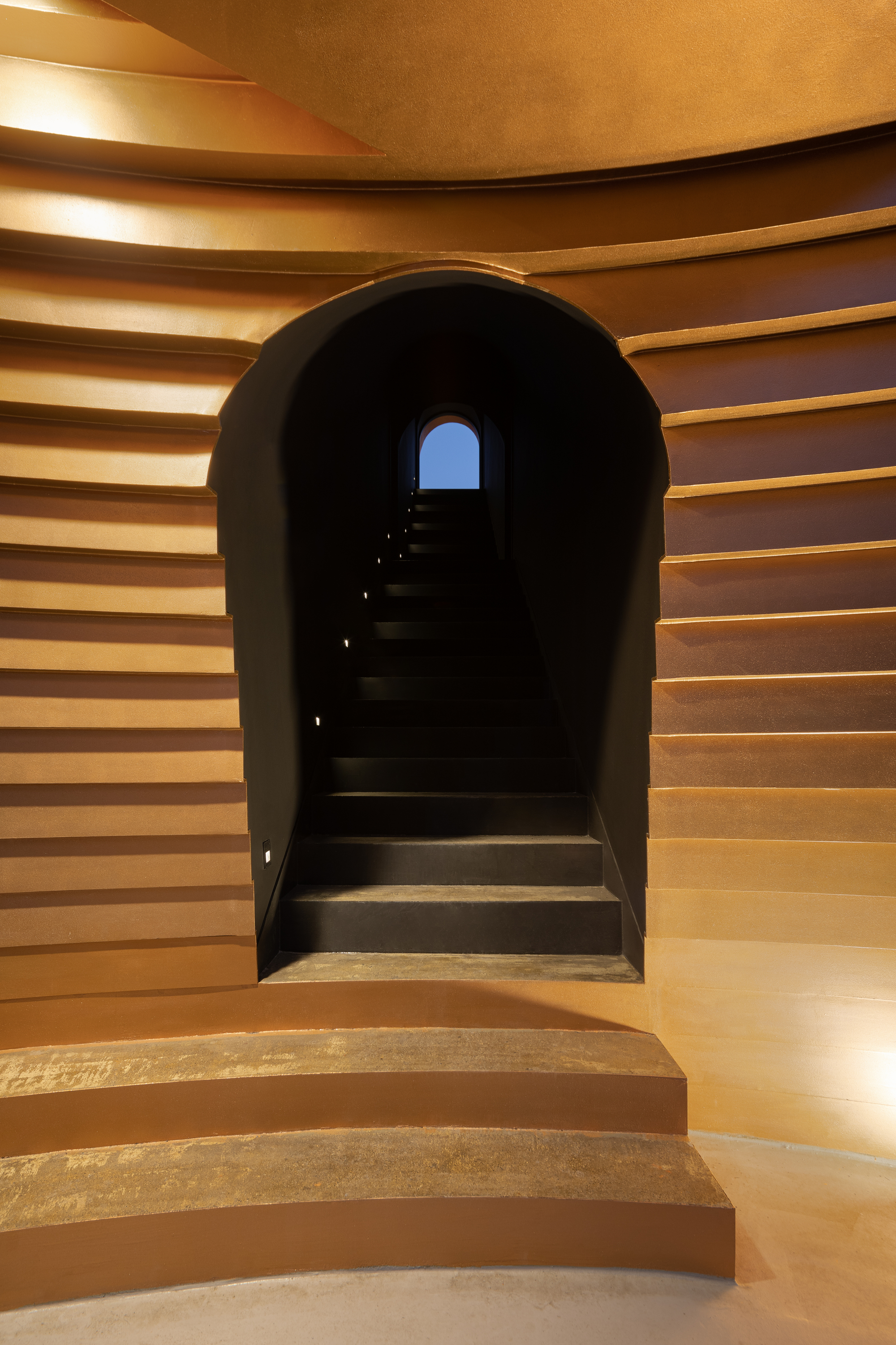 Interior view through the illuminated timber-slat tunnel showing stairs ascending toward a blue arched opening