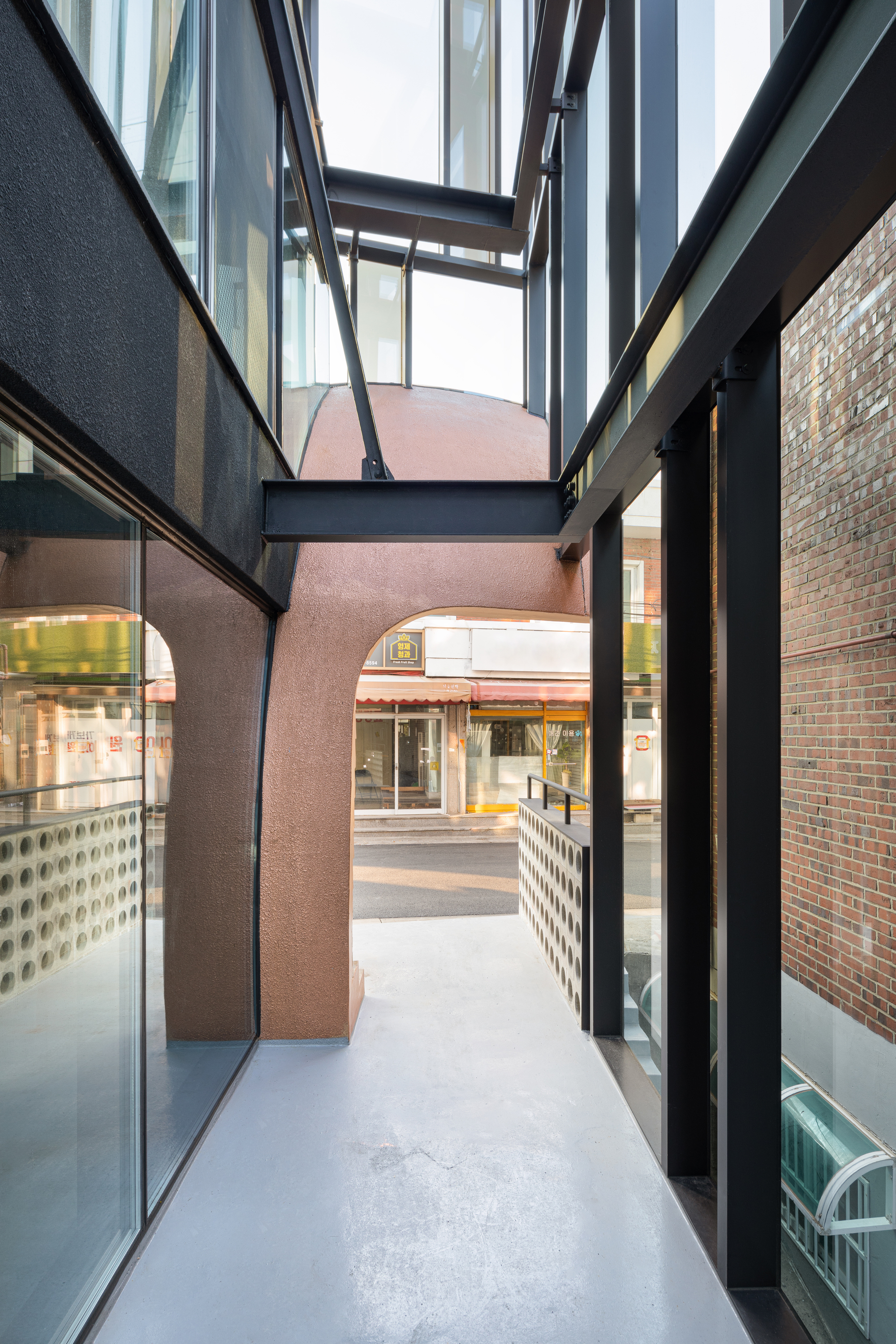 Narrow courtyard passage with glass walls, steel bridges overhead, and a pink stucco volume beyond