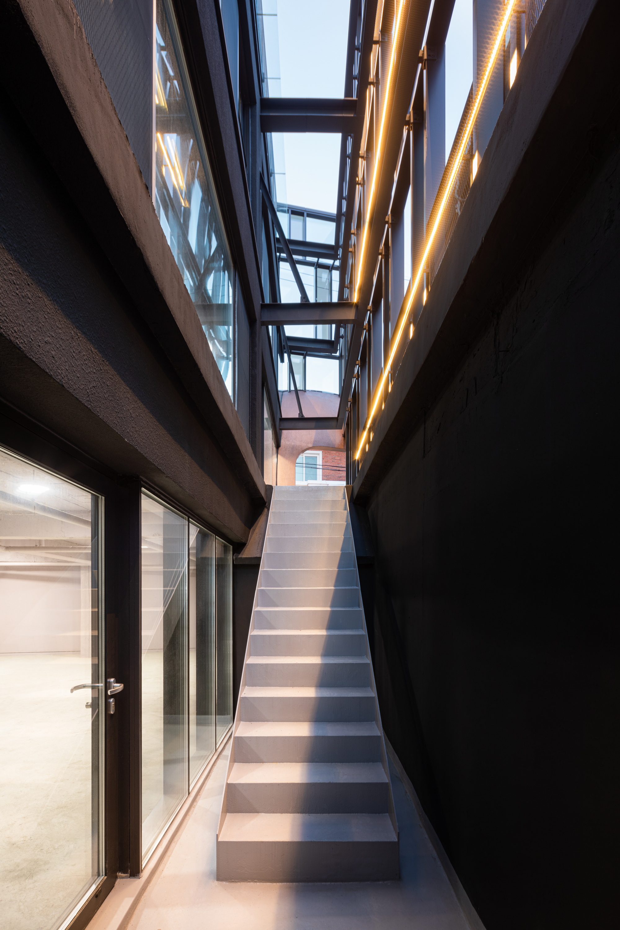 Narrow interior stair passage between black walls with steel-framed bridges and warm strip lighting at dusk