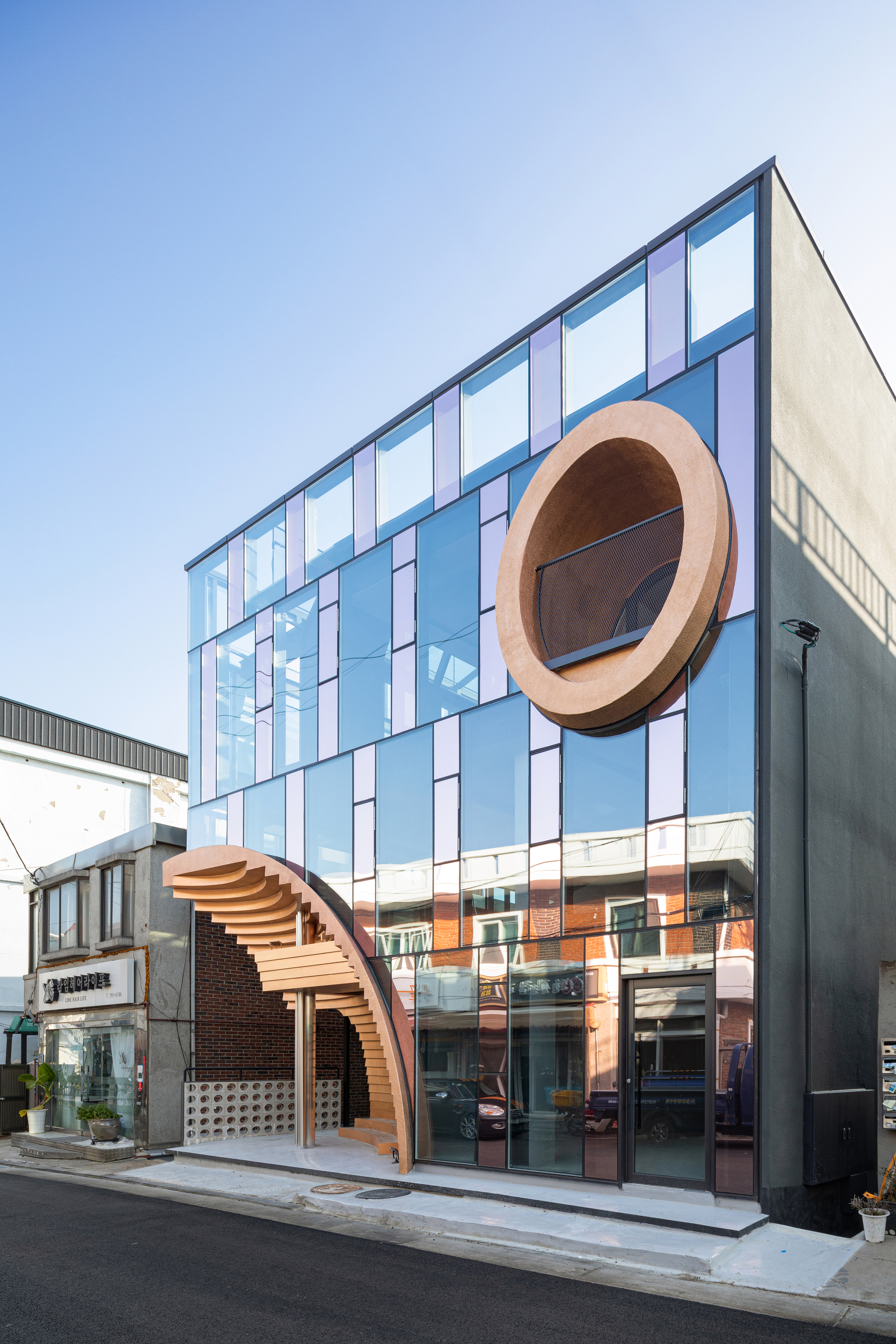 Street facade with pale blue and pink glazed panels punctuated by two laminated timber arches at ground level