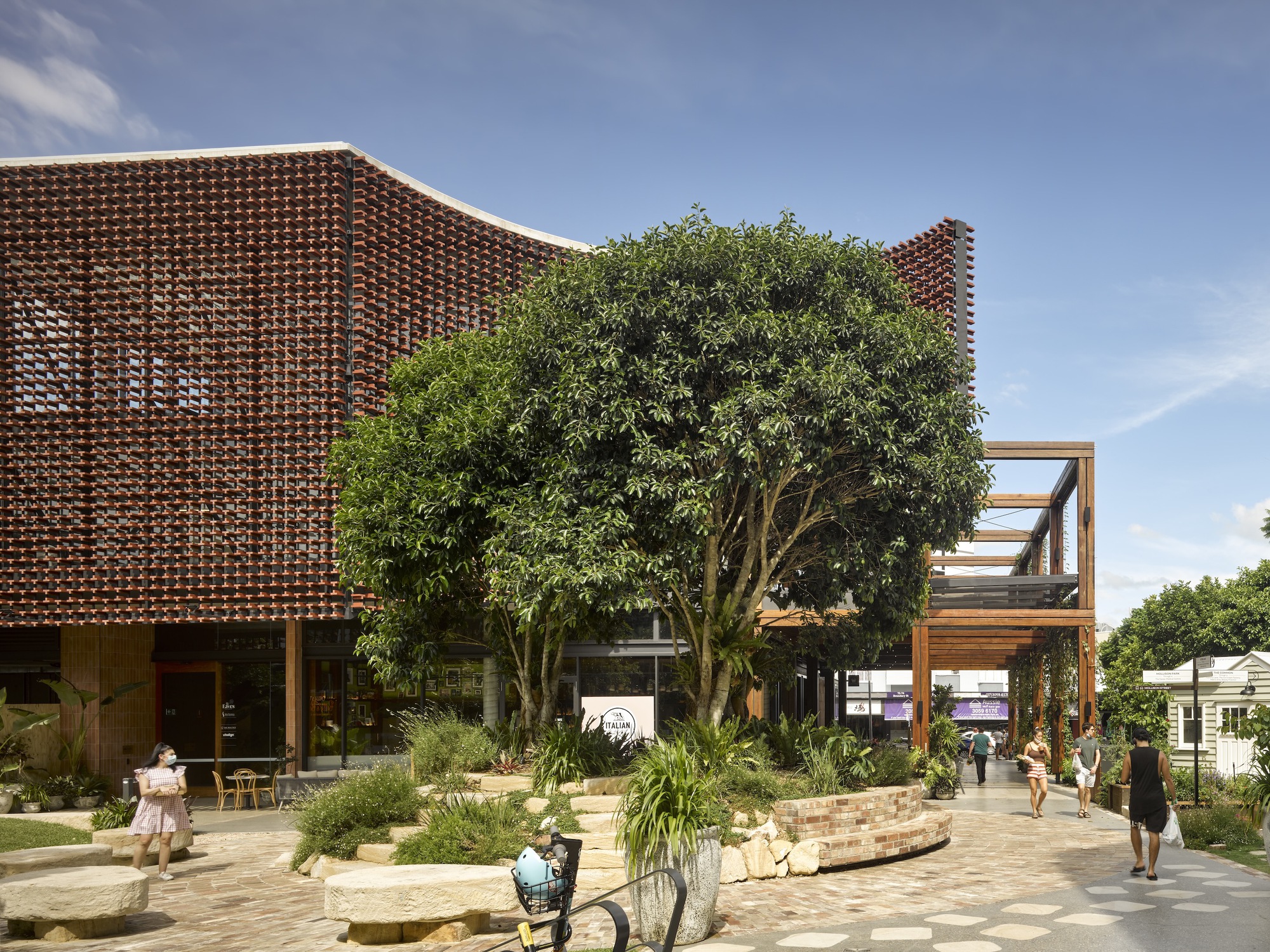Perforated terracotta brick screen wrapping the facade above a paved plaza with visitors and planted beds