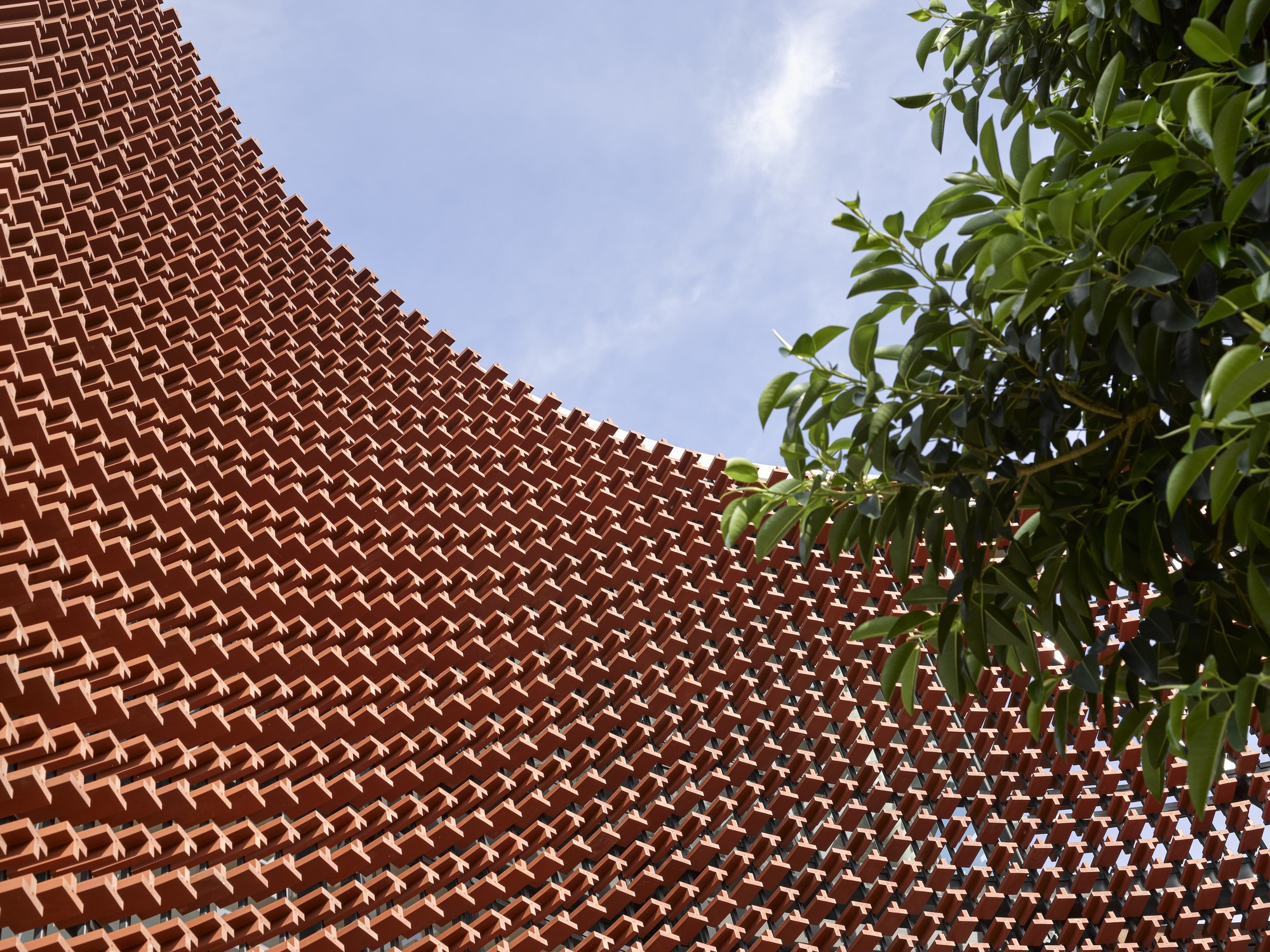 Close-up of the perforated terracotta brick pattern curving against tree foliage and blue sky