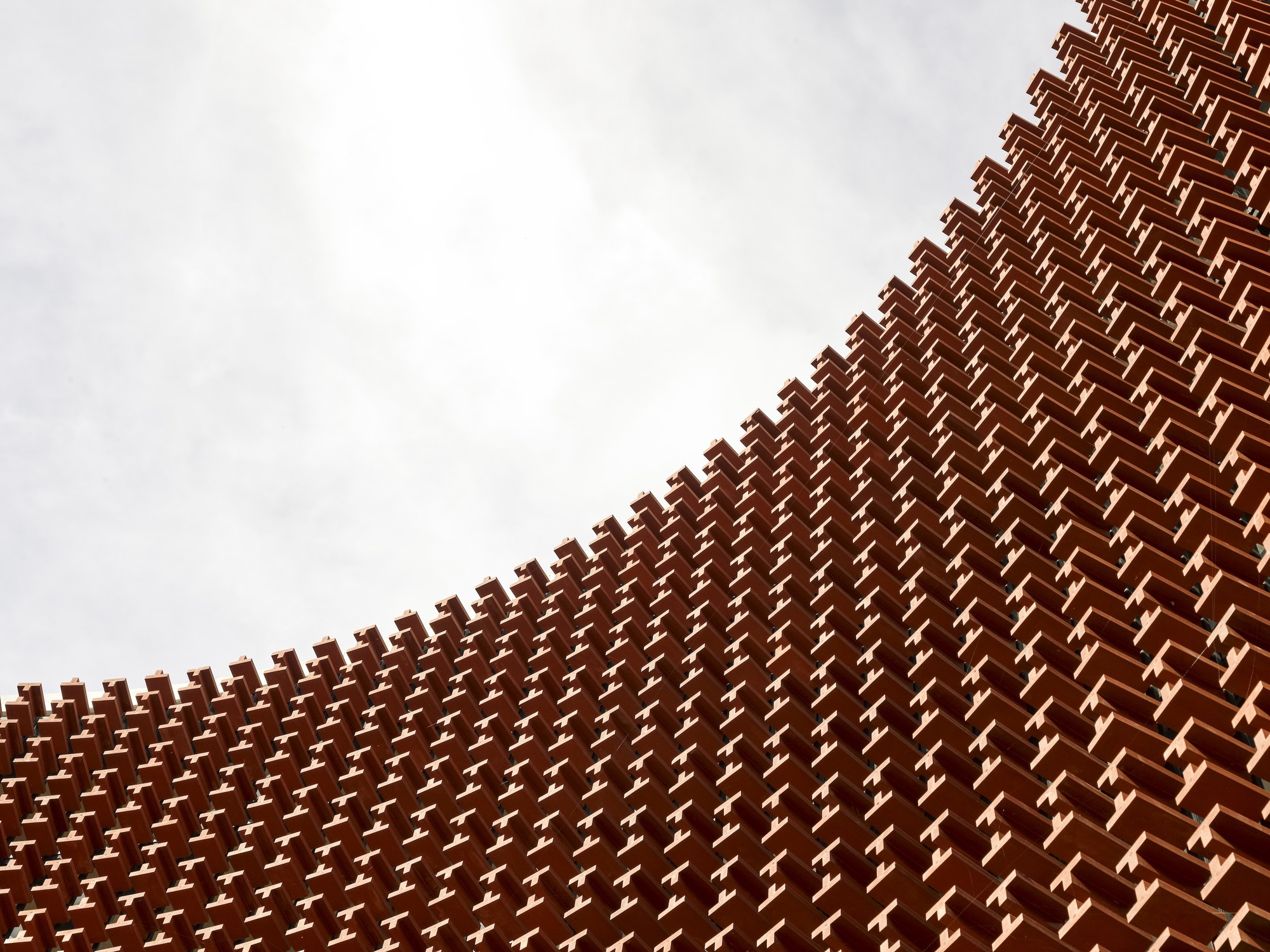Upward view of terracotta brick lattice pattern forming a diagonal facade under overcast sky