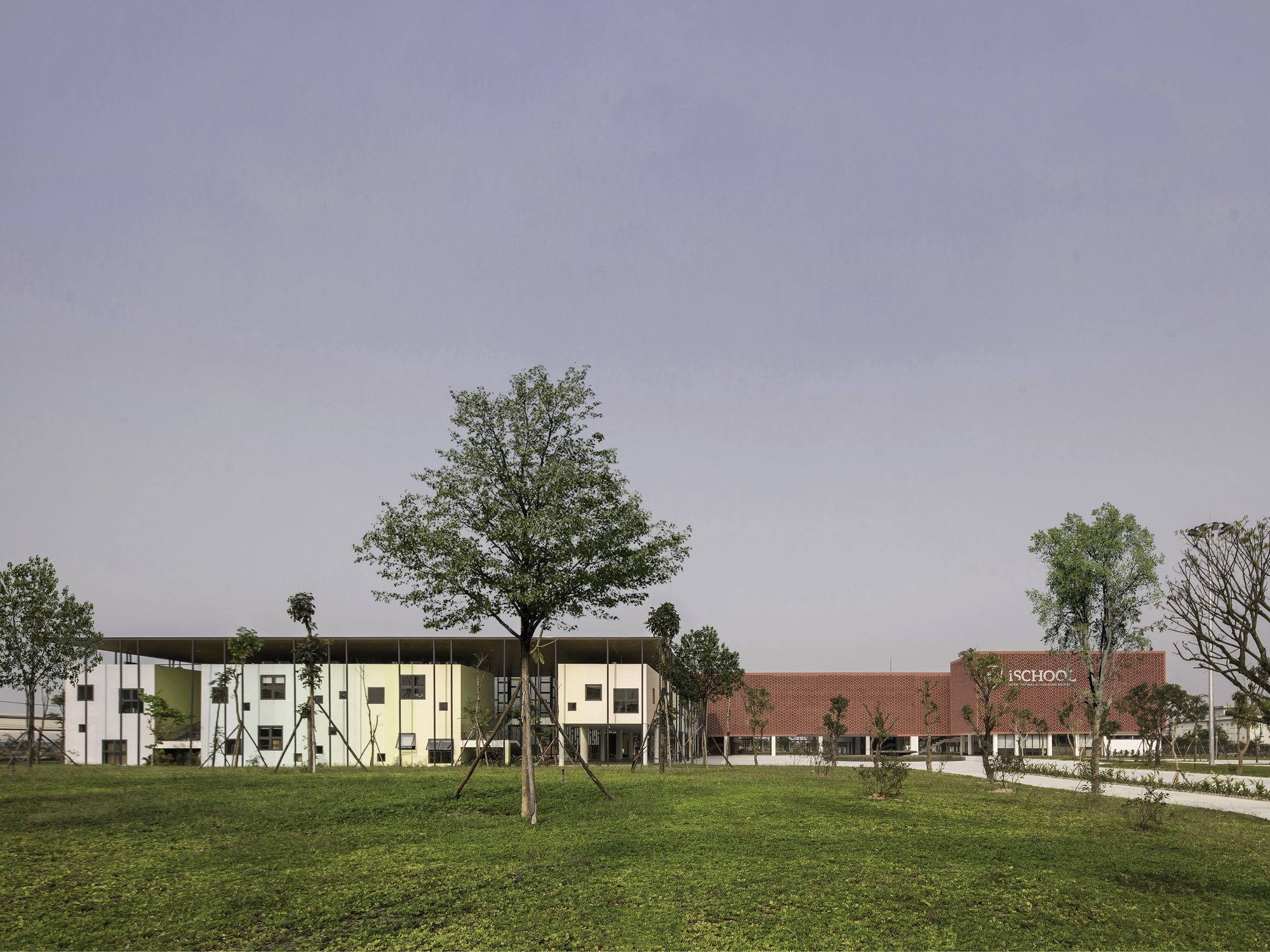 Campus view with the two-story facade and brick volume beyond a grassy field and young trees