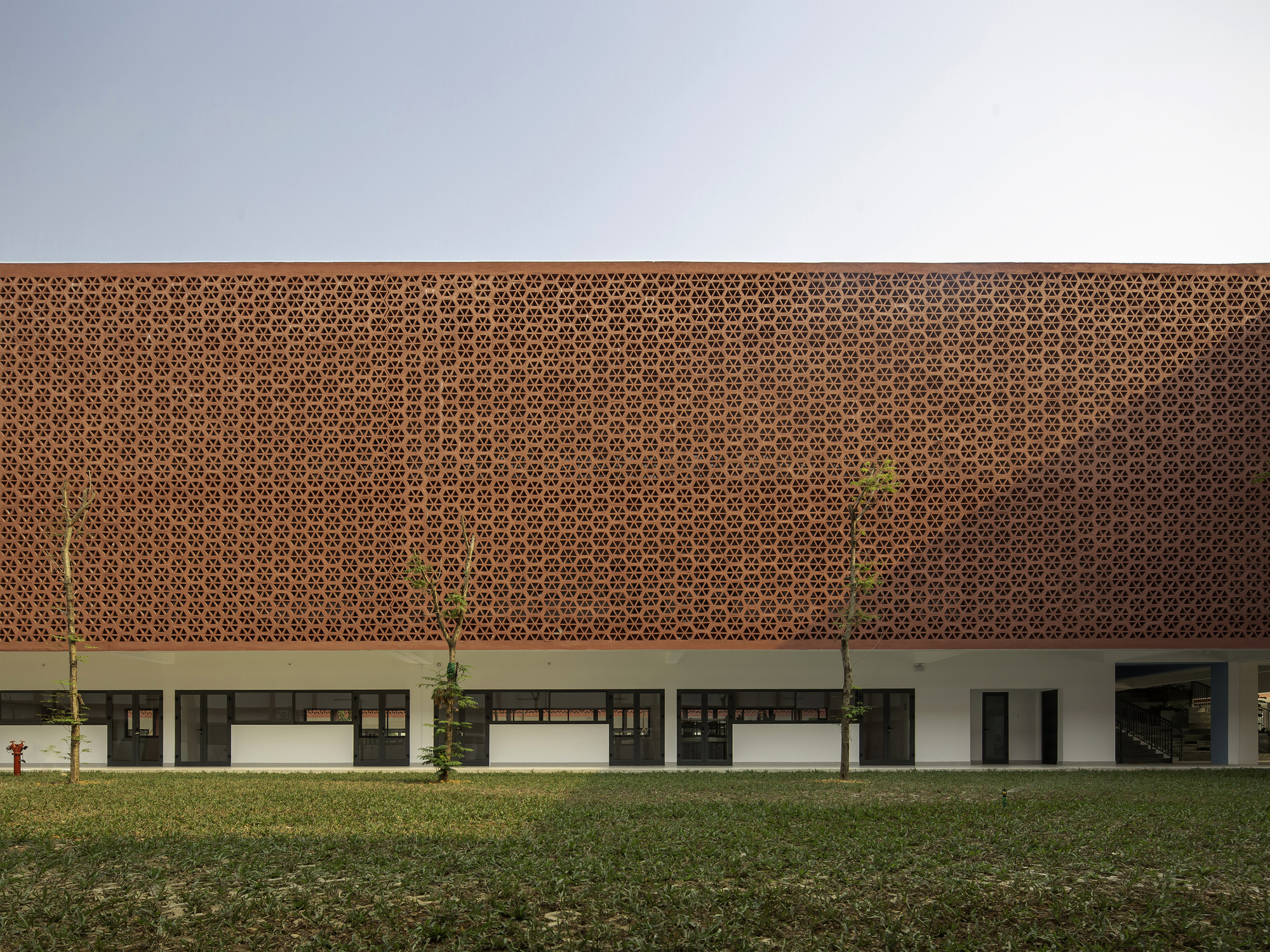 Two-story facade with perforated terracotta screen above white columns and planted saplings on grass lawn