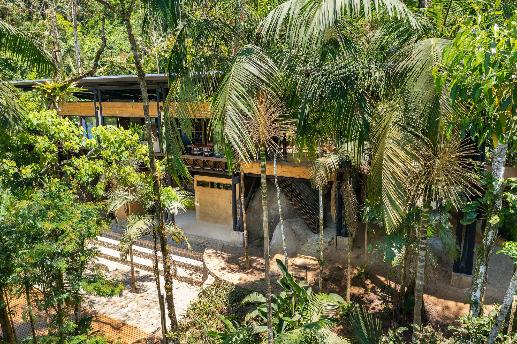 Two-story volume with timber and rammed earth walls framed by palm fronds and jungle vegetation