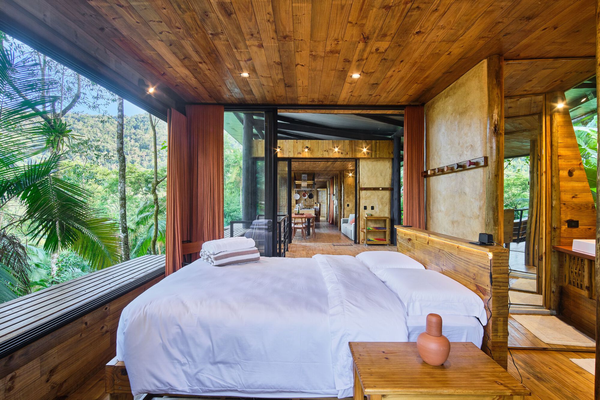 Bedroom with timber platform bed and floor-to-ceiling glazing overlooking the forested slope