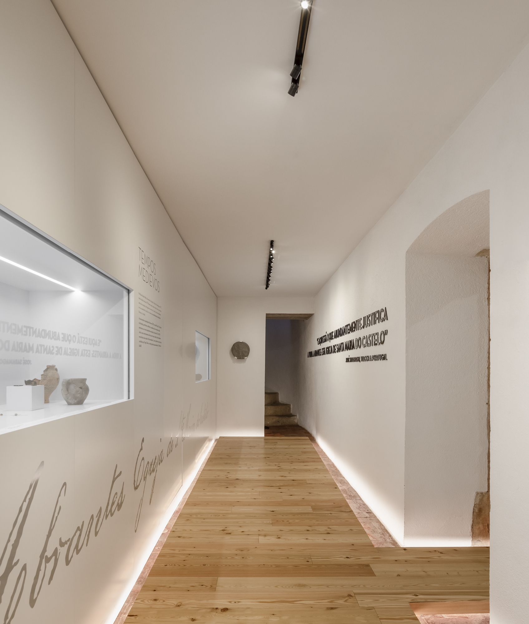 Gallery corridor with timber flooring, white walls, linear floor lighting, and display niches containing artifacts