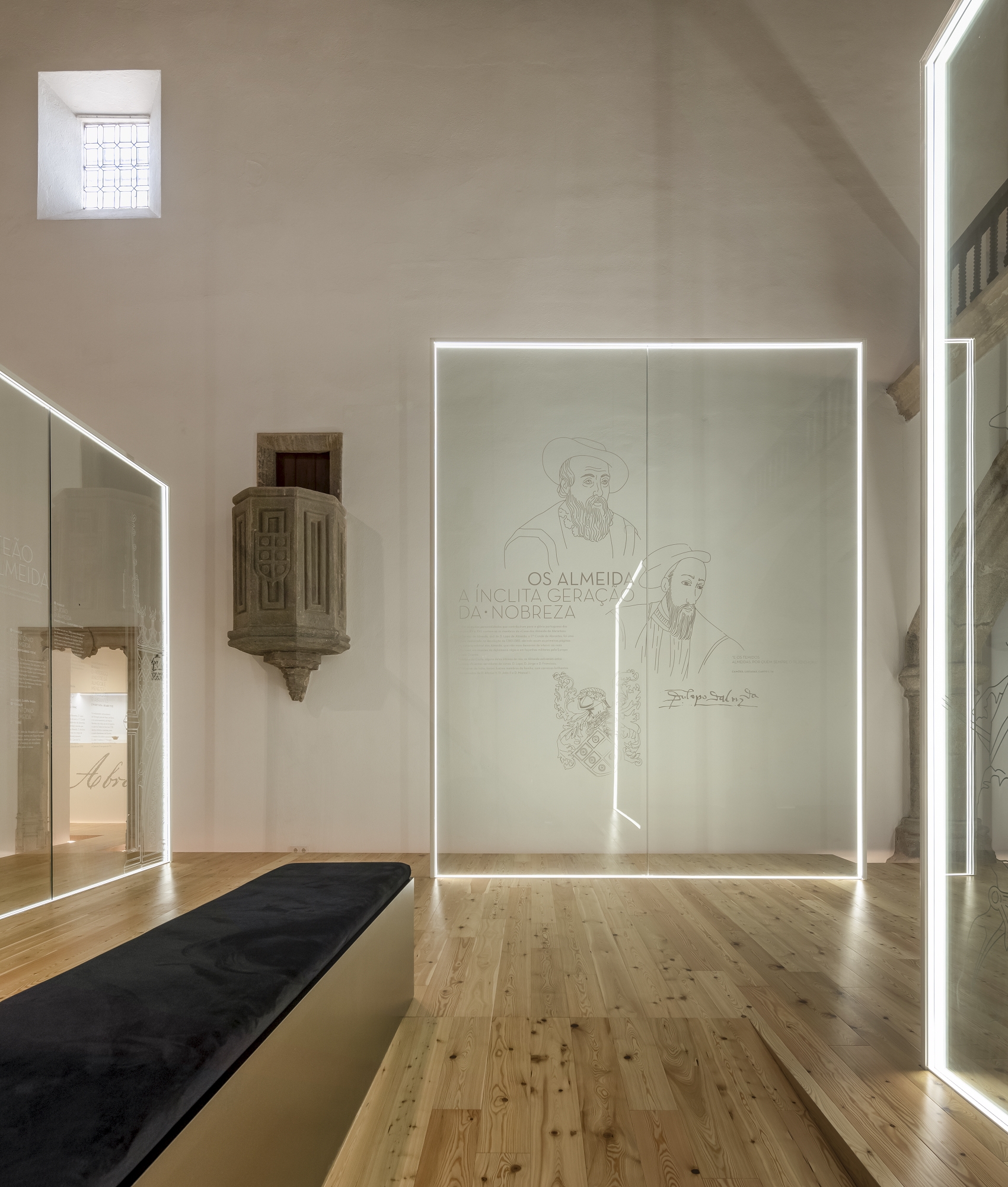 Exhibition room with vaulted ceiling, illuminated glass panels featuring line drawings, and historic stone carved element