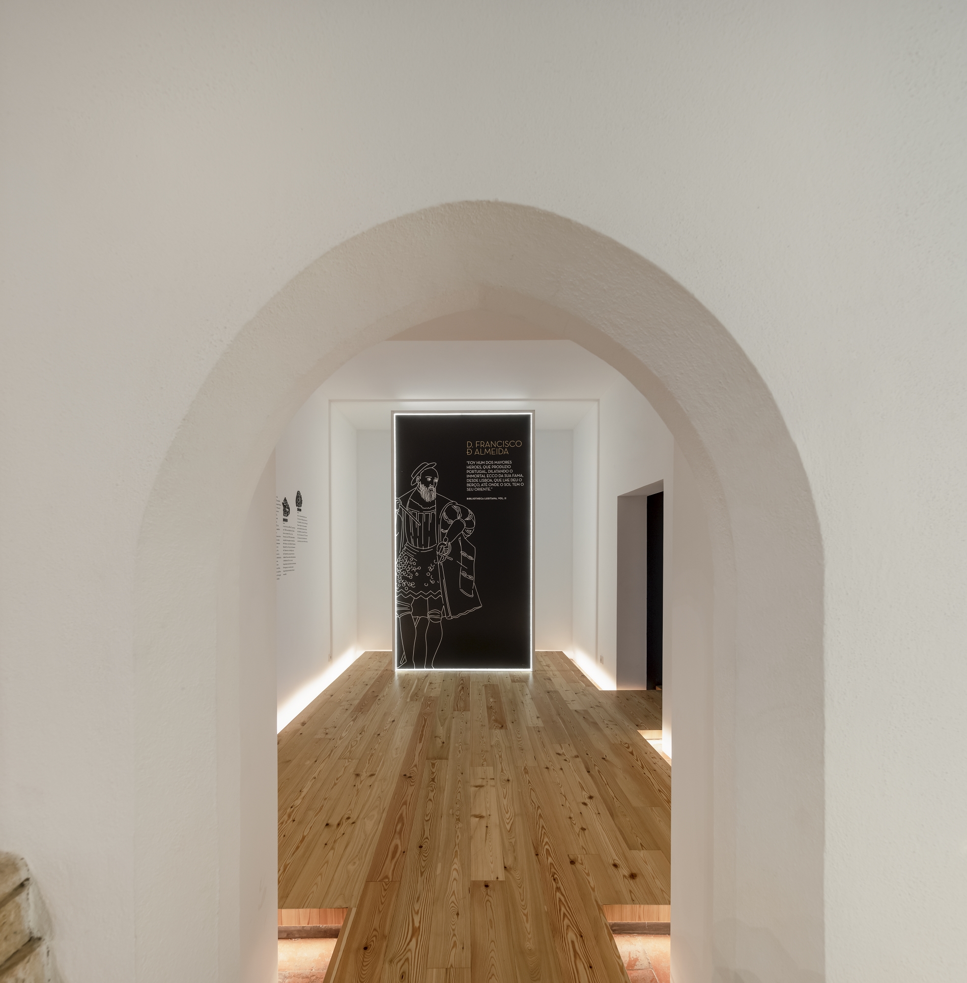View through whitewashed stone arch toward gallery space with backlit graphic panel on far wall