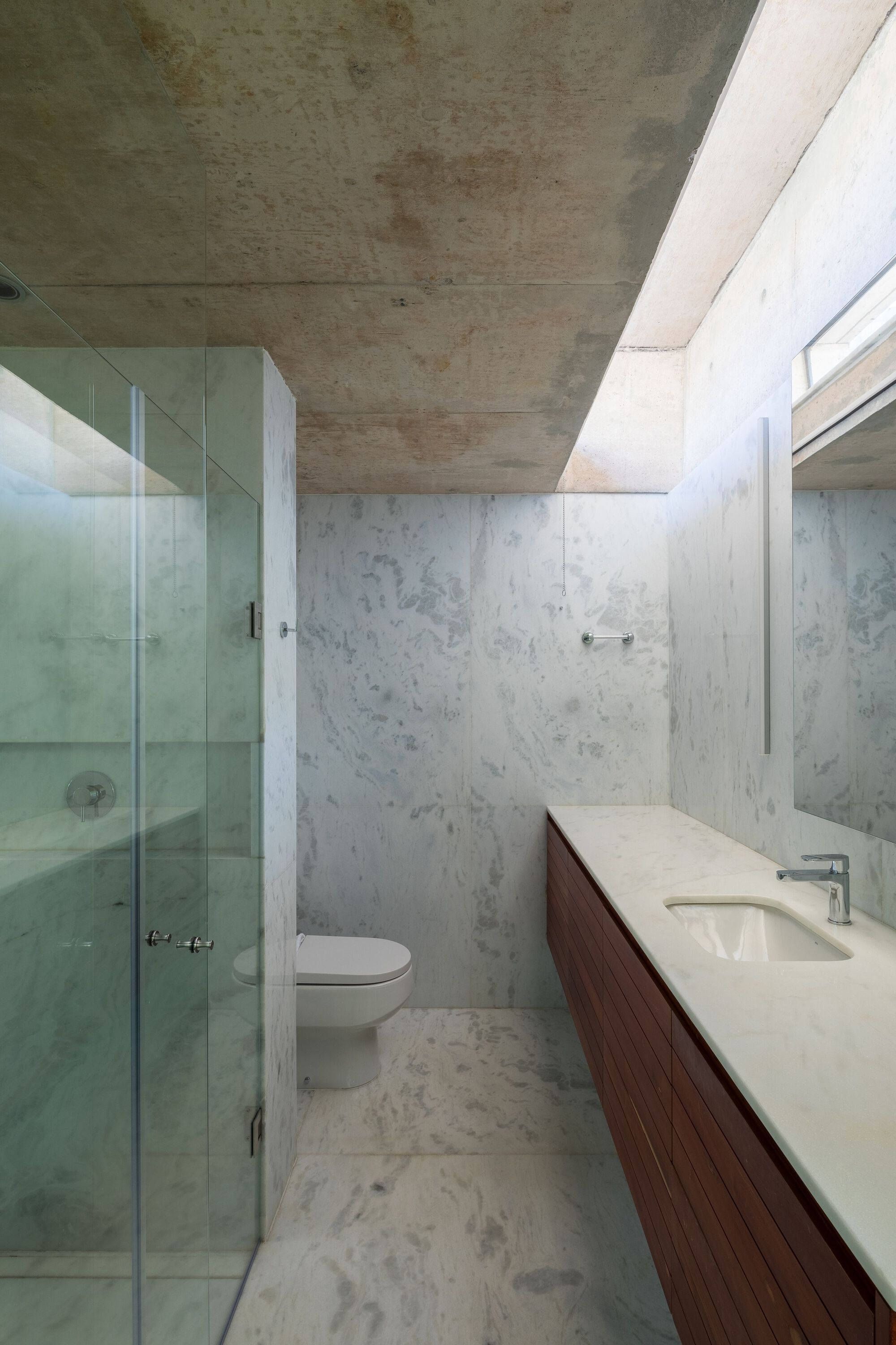 Bathroom with exposed concrete ceiling, walls and floor, glass shower enclosure and walnut vanity cabinet