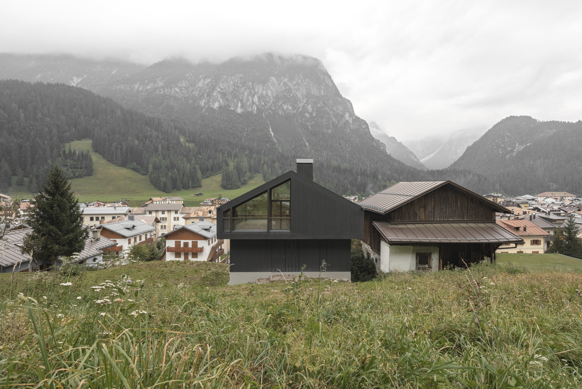 Two-part house with dark cladding and glass volume beside weathered barn under cloudy mountain sky