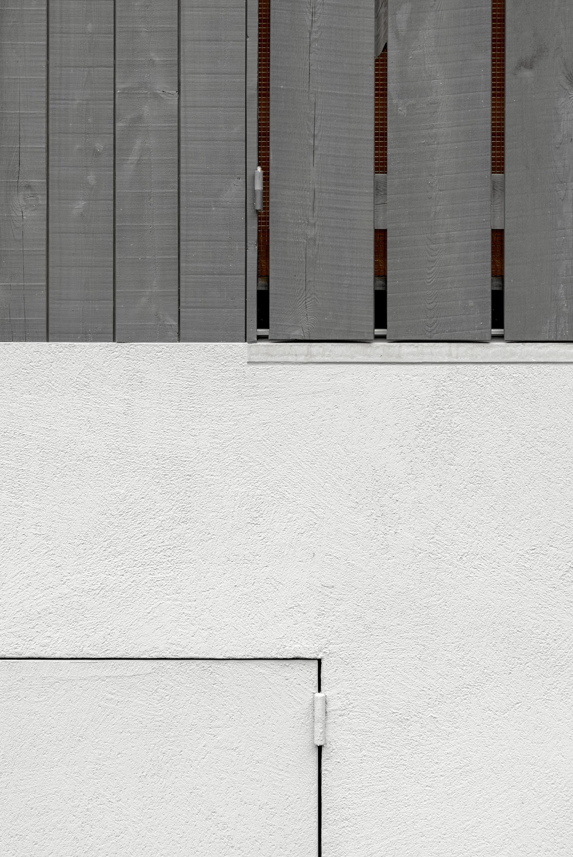Concrete facade with vertical timber-lined recesses above a white stucco wall and paved courtyard