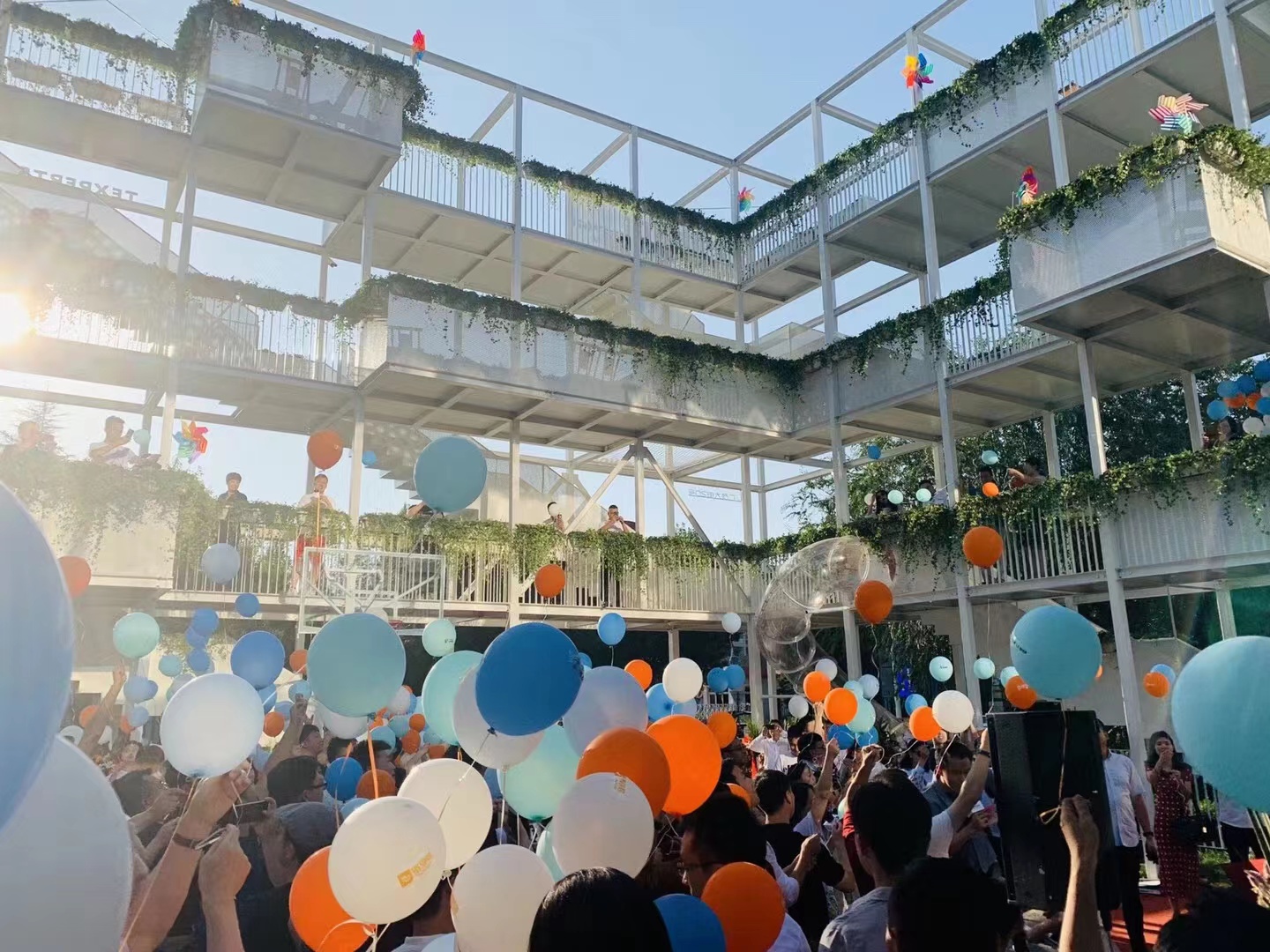 Upward view through released balloons toward planted balconies with white railings in afternoon sunlight