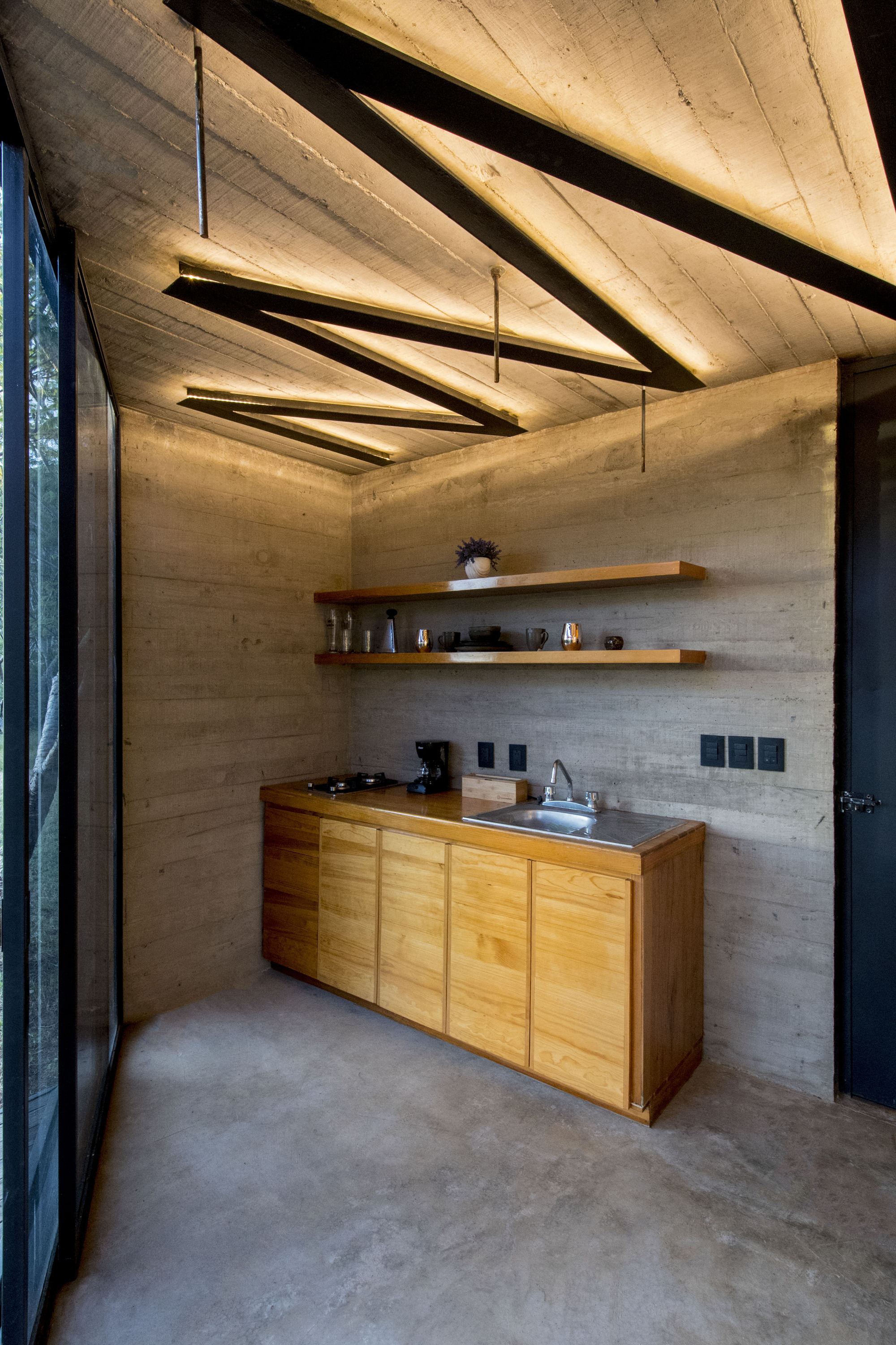Compact kitchen with timber cabinetry beneath triangular suspended light fixtures and board-formed concrete walls
