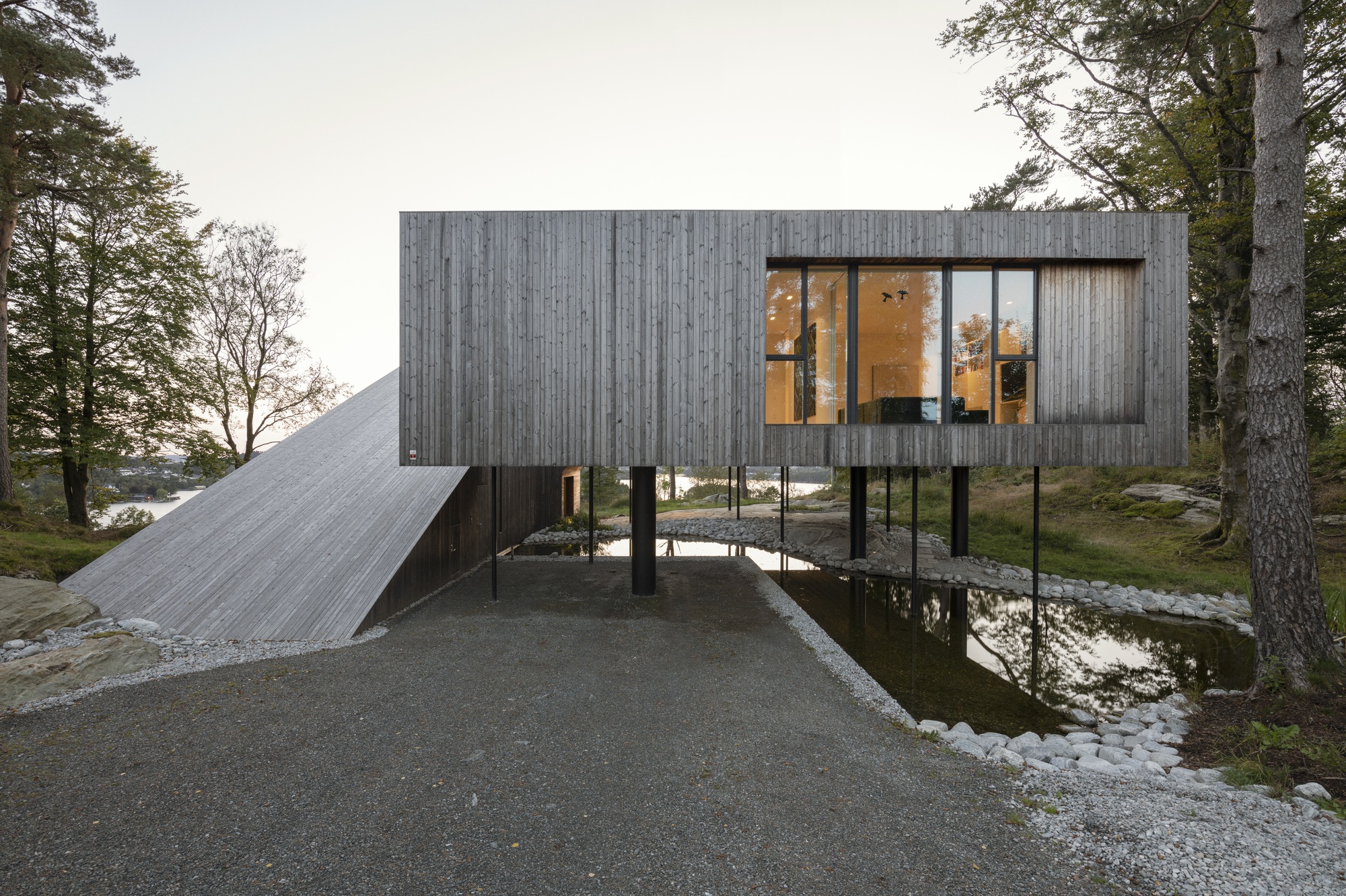 Entry view of the elevated timber box and sloped roof with illuminated interior at dusk