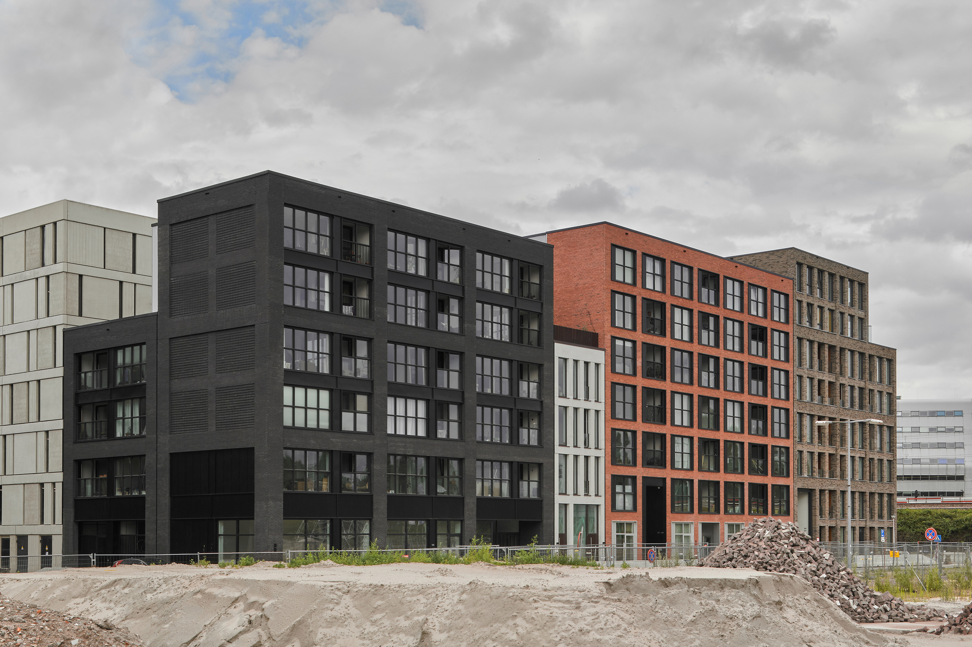 Street view of three multiunit blocks with orange brick, black cladding and grey panel facades across undeveloped ground