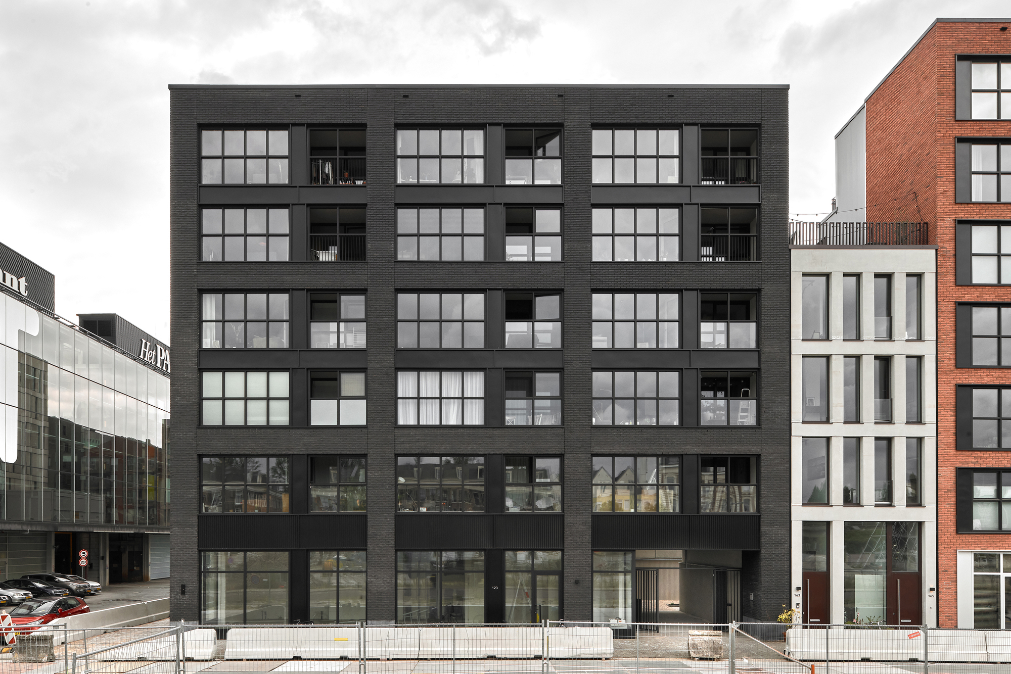 Black cladded facade with large gridded windows and recessed balconies flanked by neighboring buildings