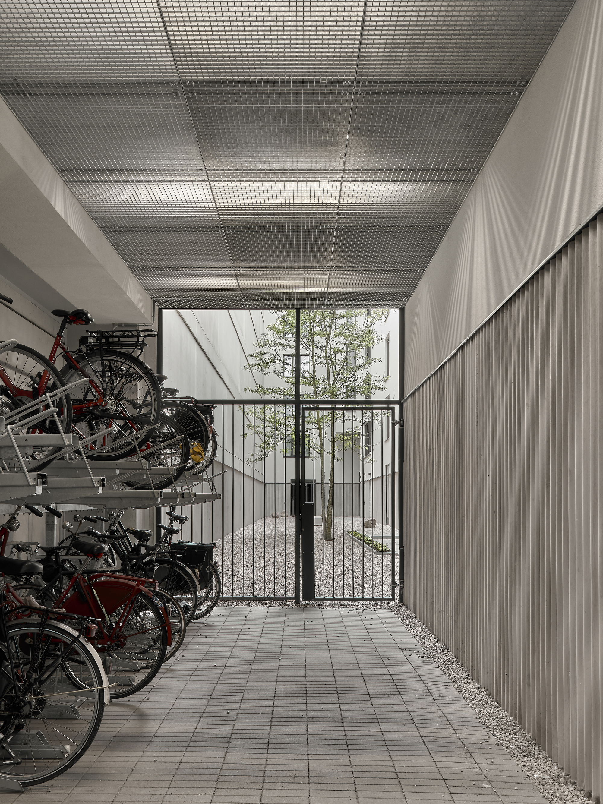 Covered bicycle storage with metal mesh ceiling and glazed gate opening to trees beyond