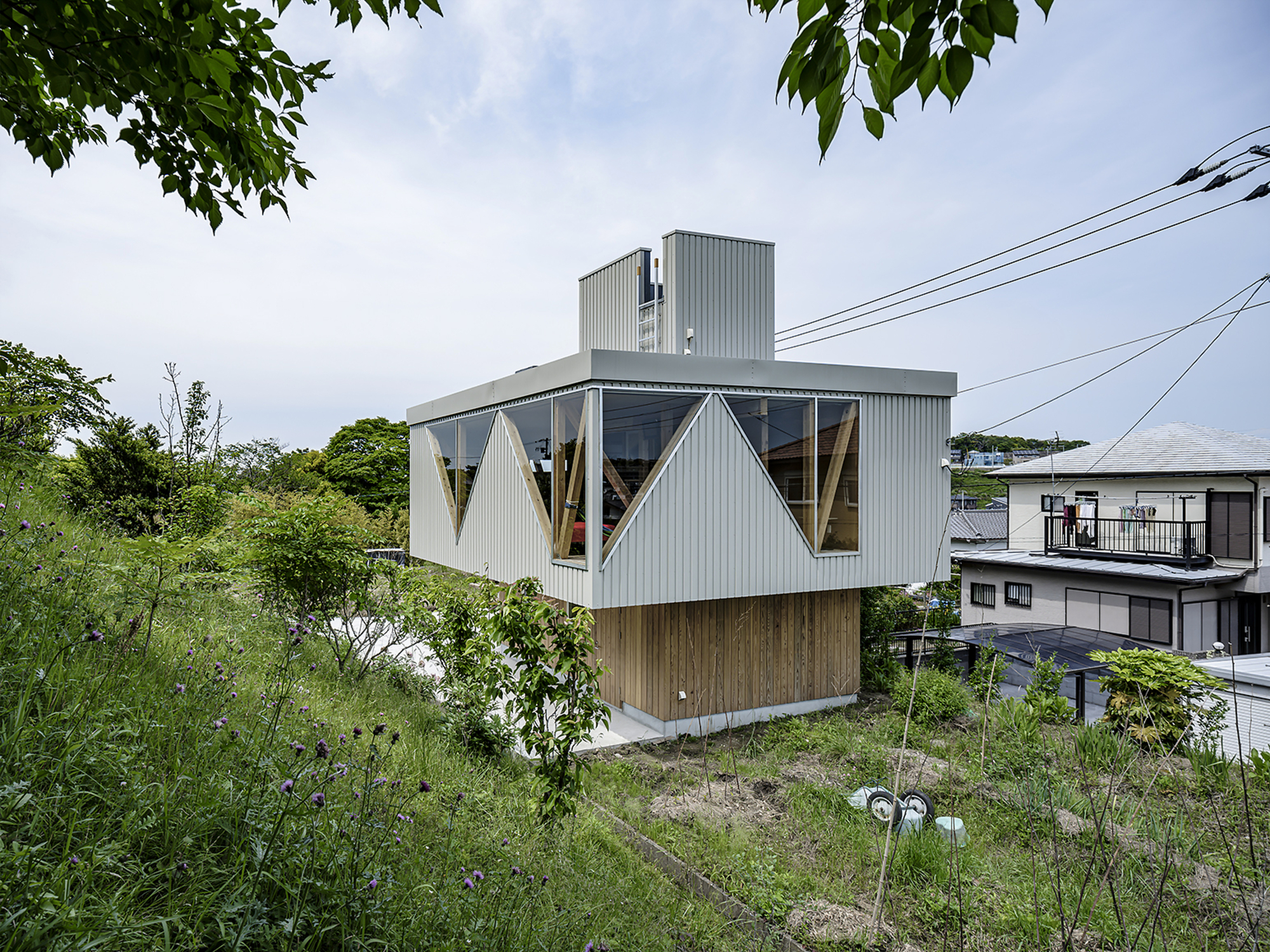 Corrugated metal facade with triangular windows set above a timber base surrounded by green vegetation
