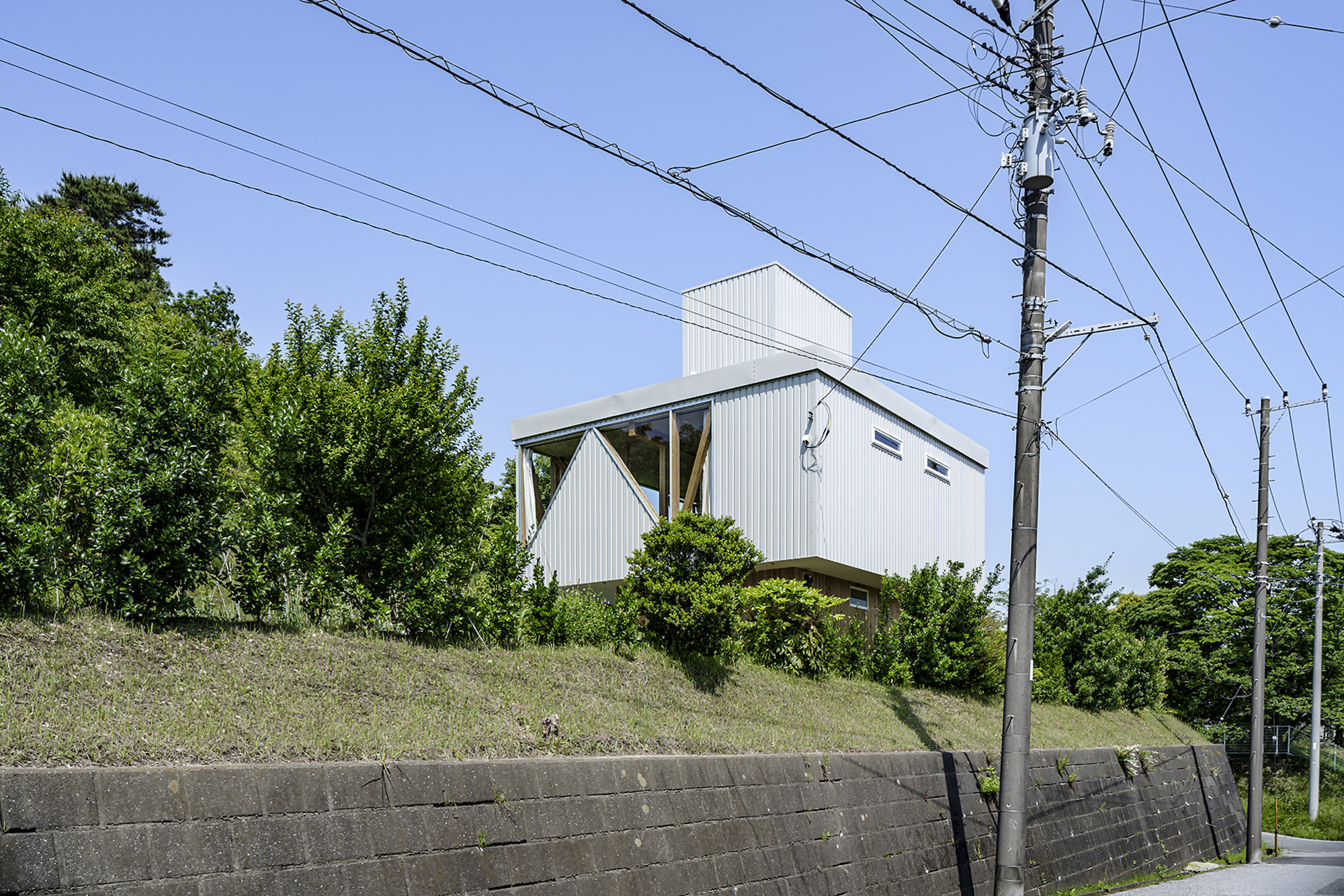 White corrugated metal house on a grassy slope with overhead power lines and concrete retaining wall