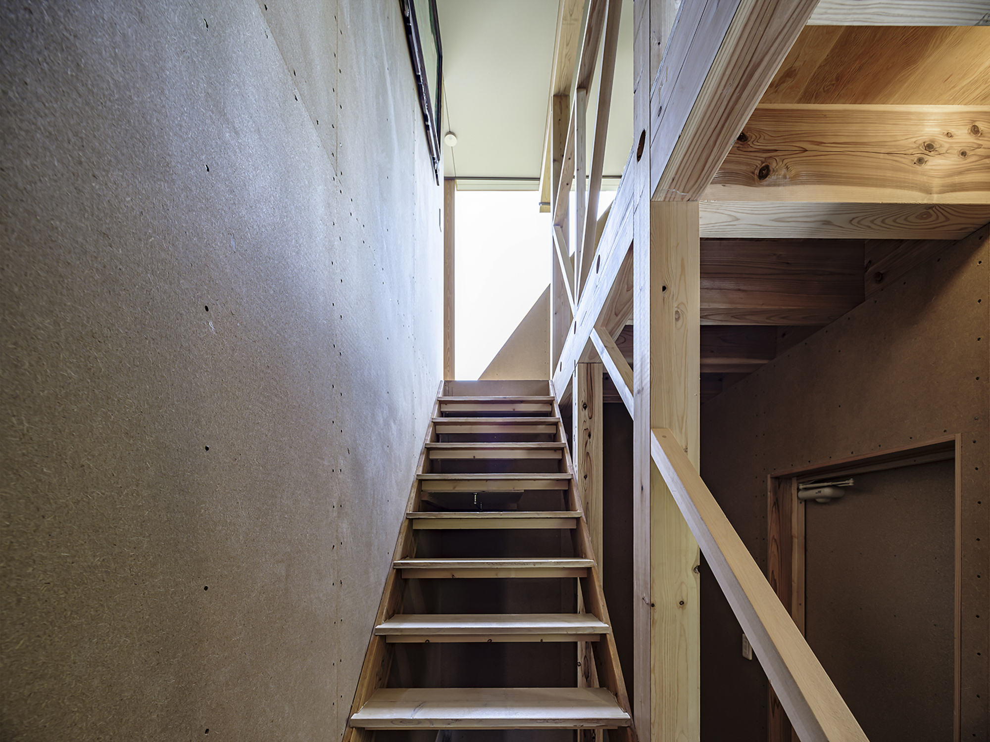 Timber stair ascending toward a bright skylight between concrete wall and cross-braced wooden railing