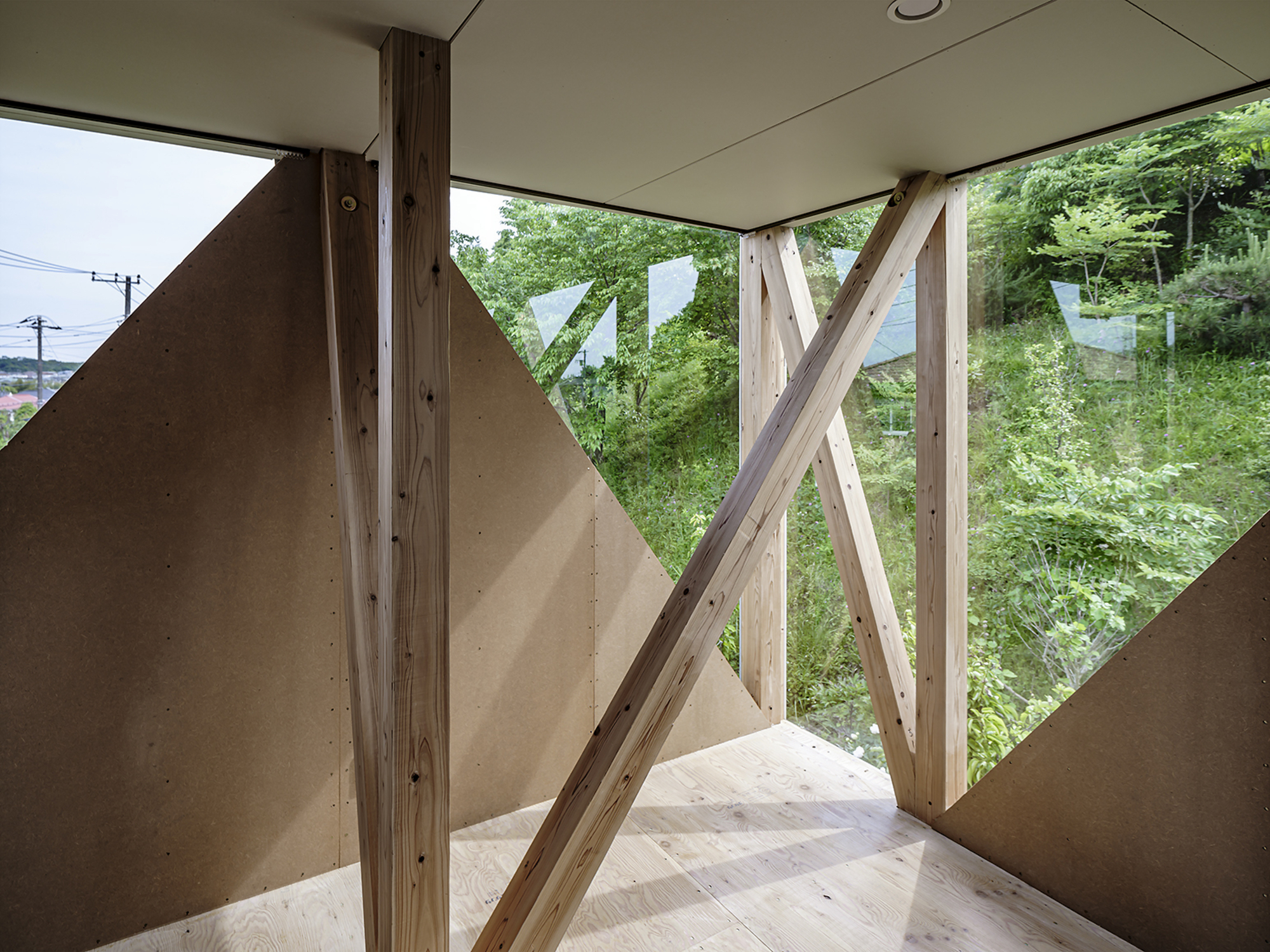 Interior corner with diagonal timber bracing framing a view of dense green foliage outside