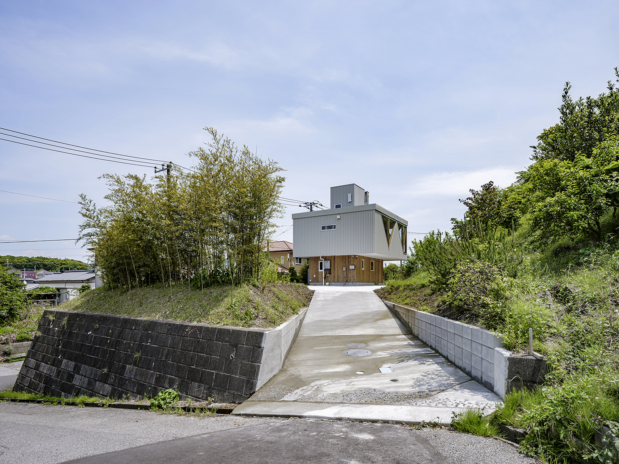 Concrete driveway rising toward a metal-clad house nestled between a wooded hillside and neighboring rooftops