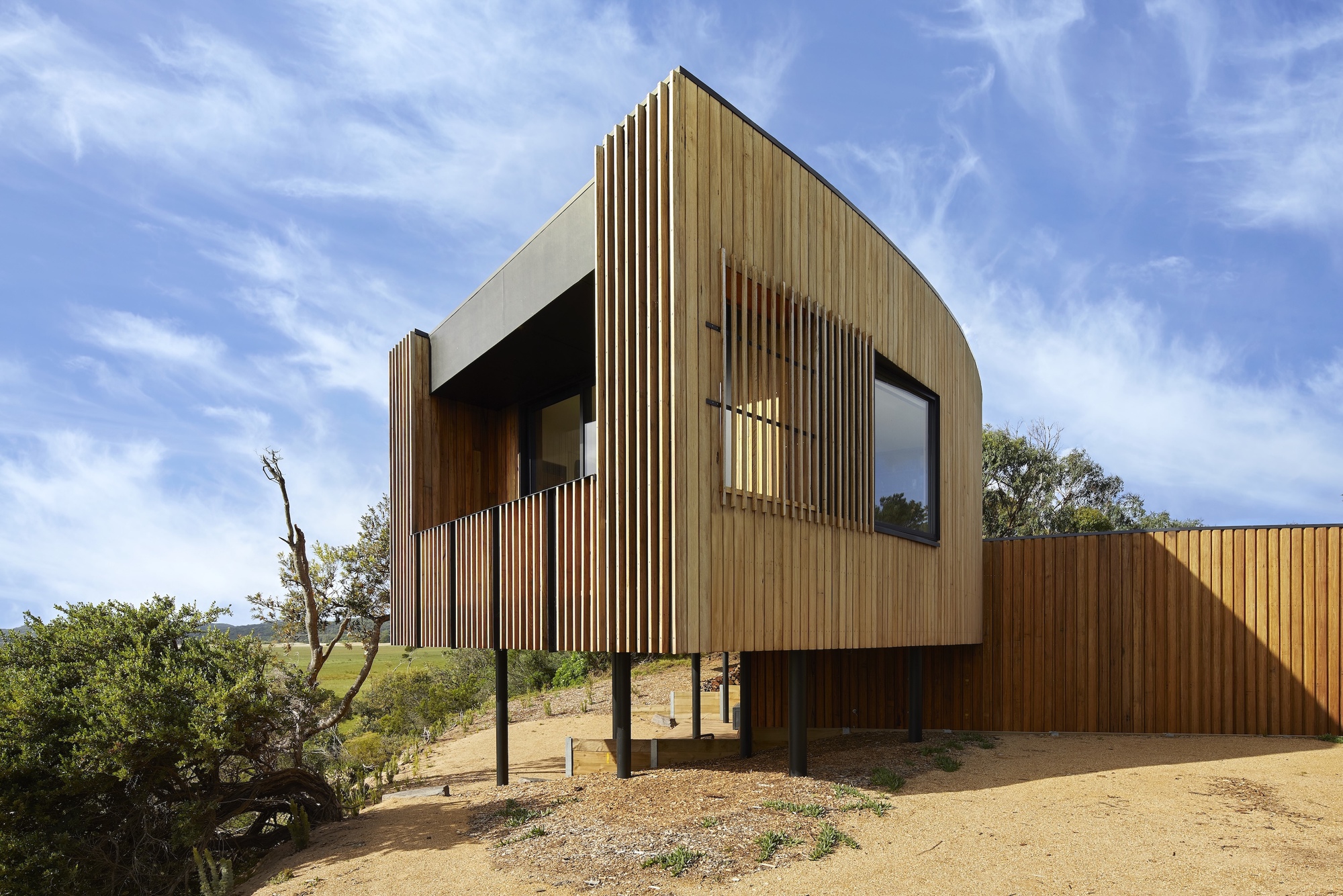 Timber-clad elevated volume with vertical slat screen and recessed openings amid coastal scrubland