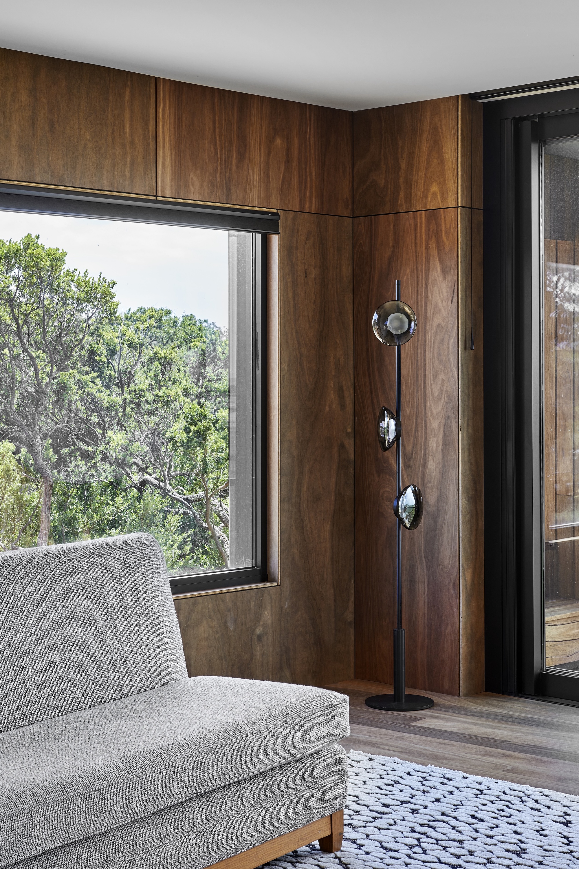 Living room corner with timber-paneled walls and black-framed window framing native trees