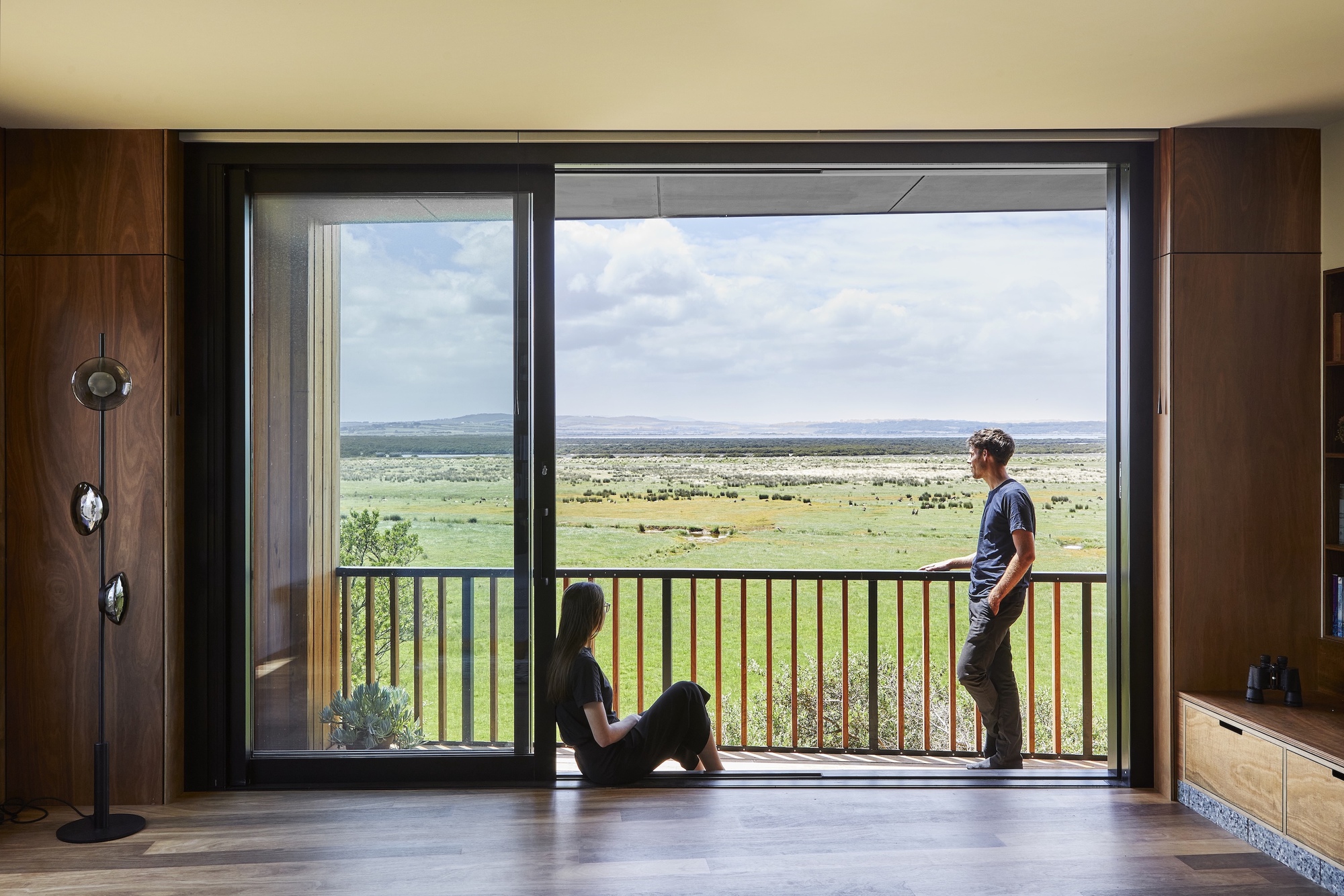 Open glass door with two figures on the threshold framing a vast grassy plain