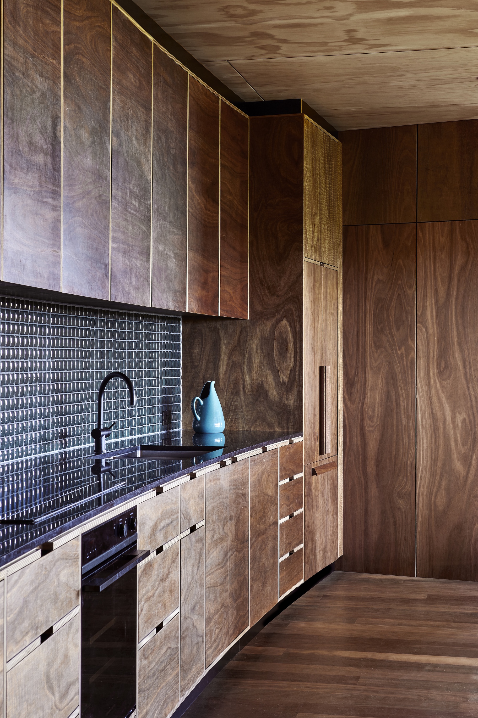 Galley kitchen with textured black tile backsplash and timber cabinetry under plywood ceiling