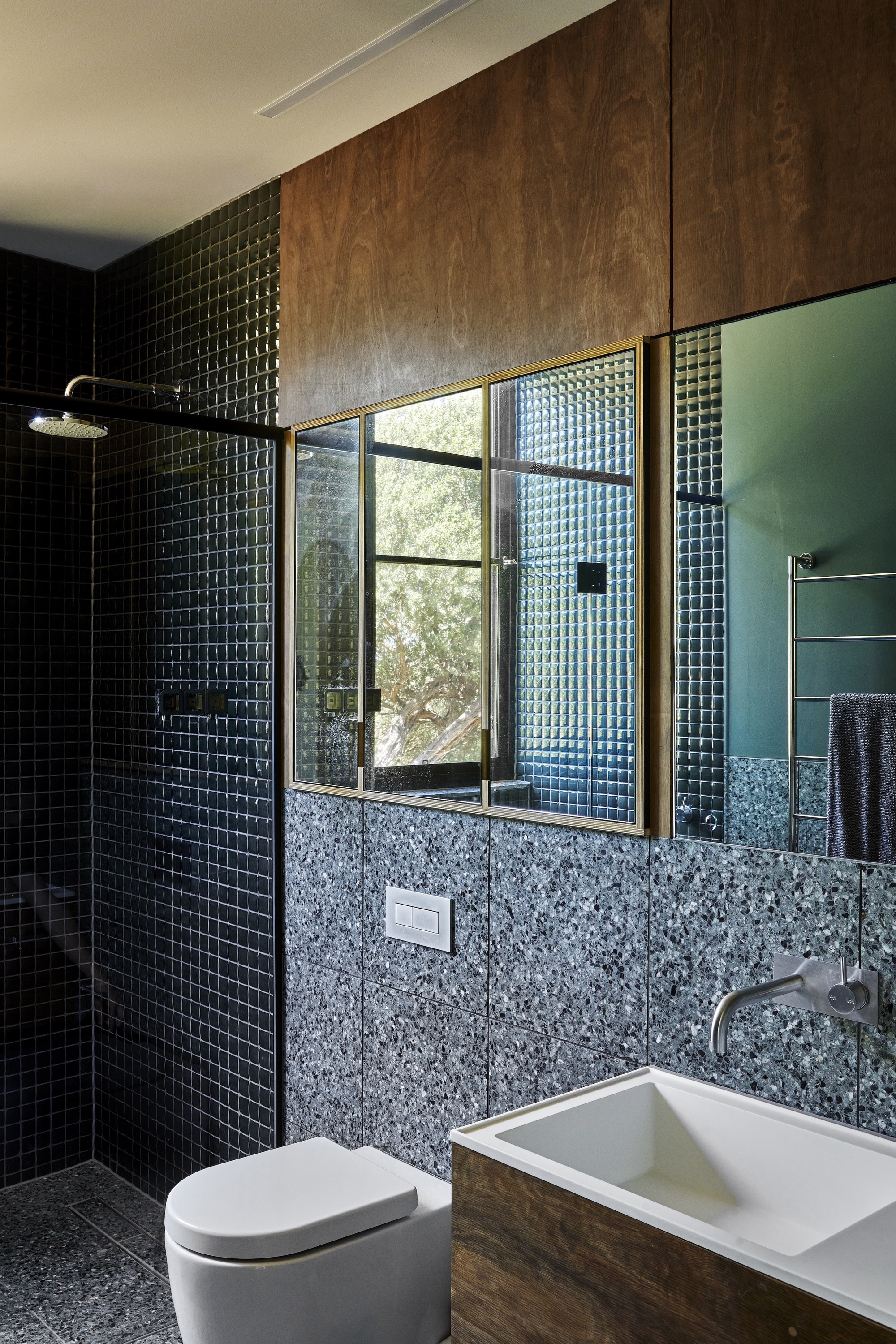 Bathroom with dark mosaic tile walls, terrazzo base, and timber-framed window to greenery outside