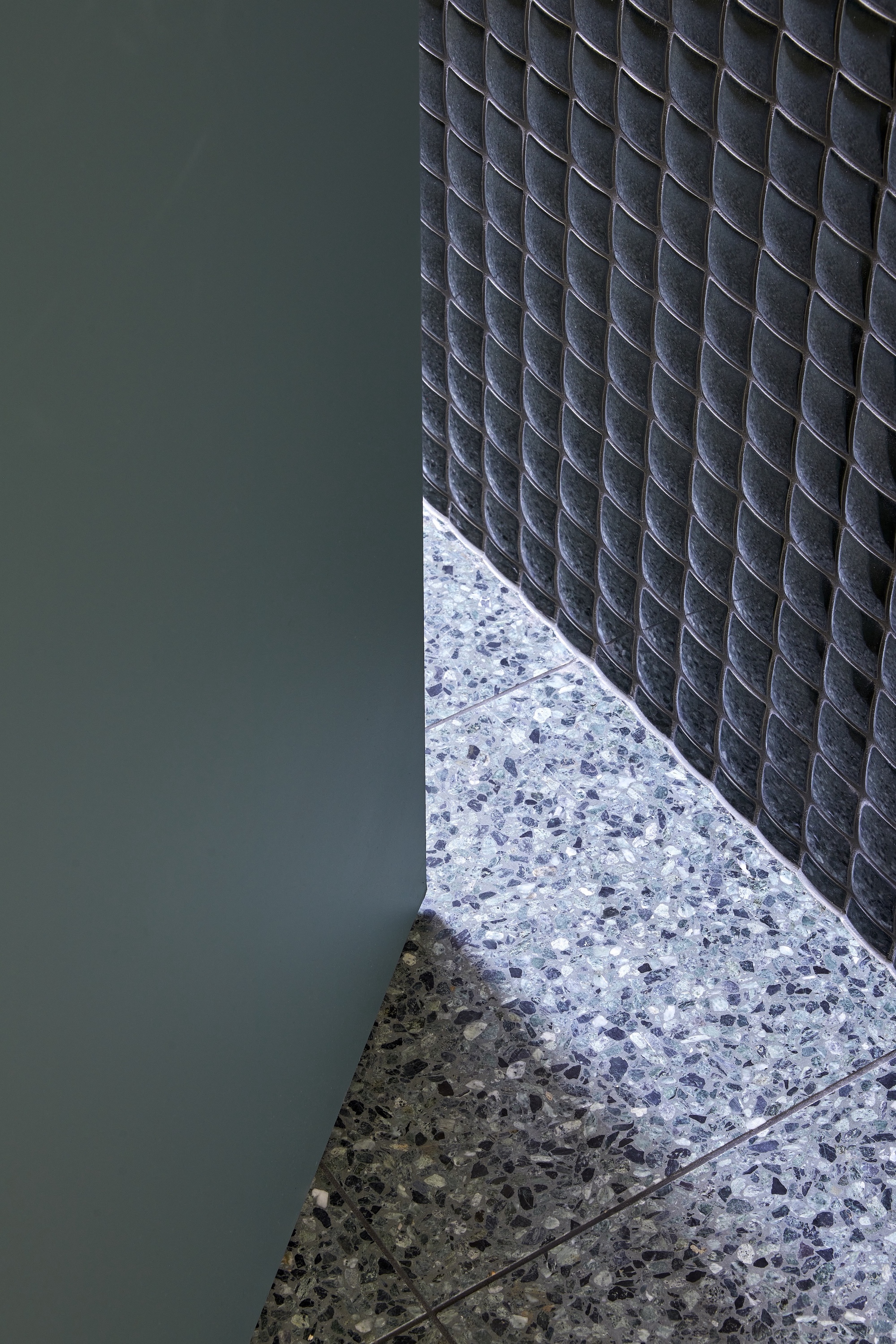 Floor detail where terrazzo meets dark mosaic tile wall with green painted surface adjacent