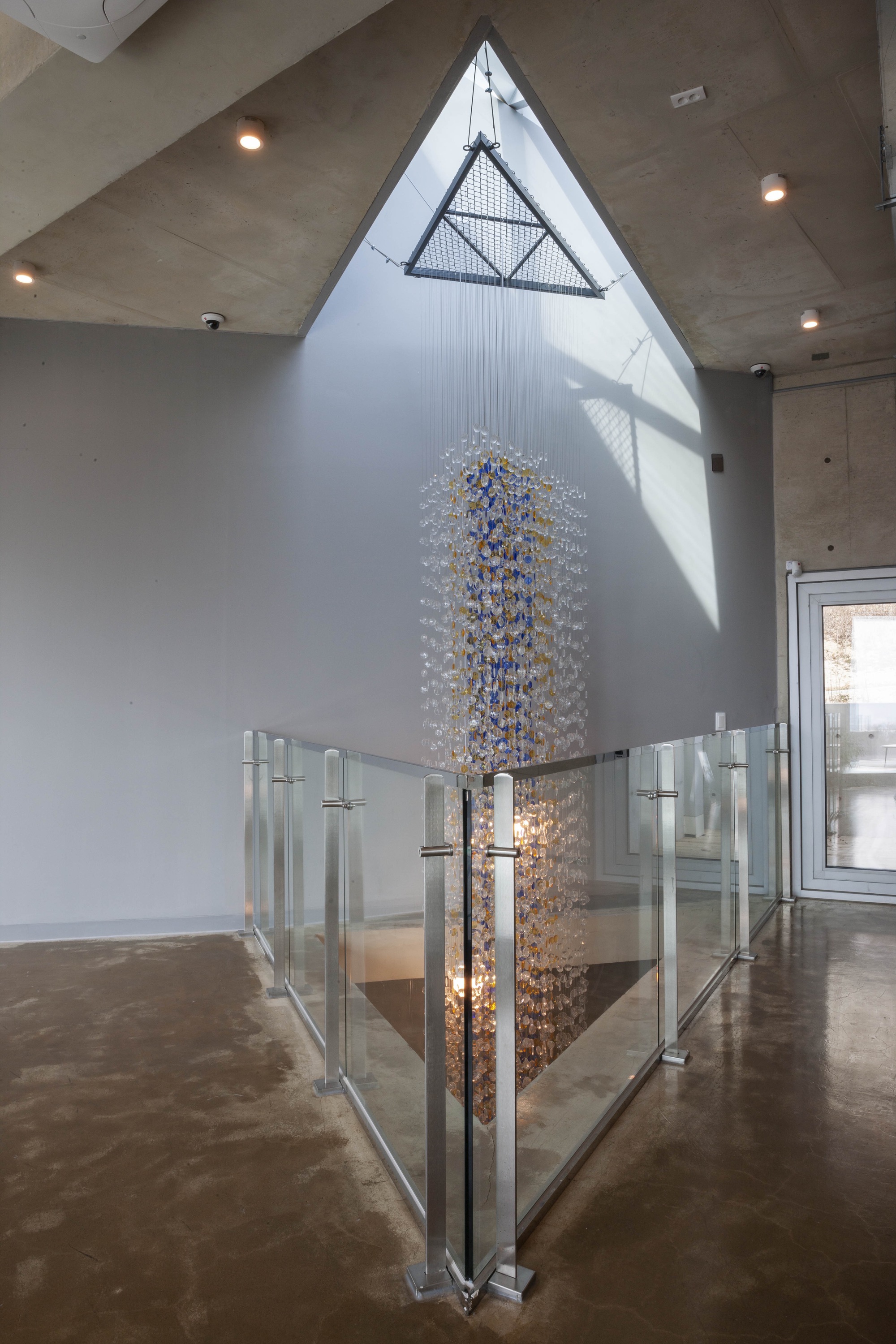 Double-height space with triangular skylight above a glass-railed stair opening and cascading chandelier
