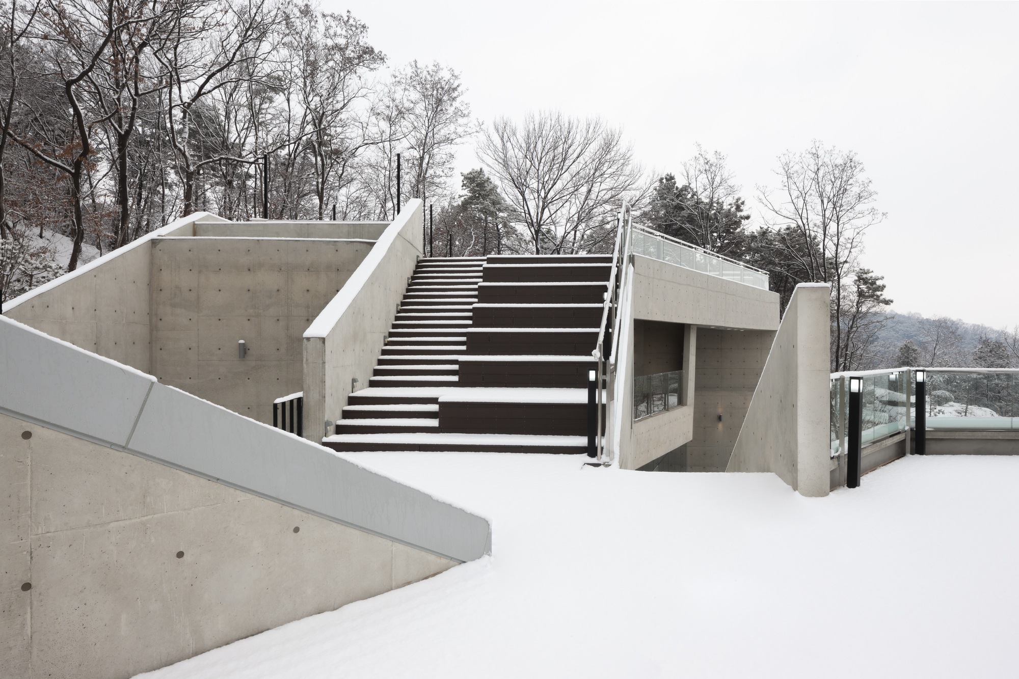 Exterior concrete stair ascending from a snow-covered terrace with bare winter trees in the background