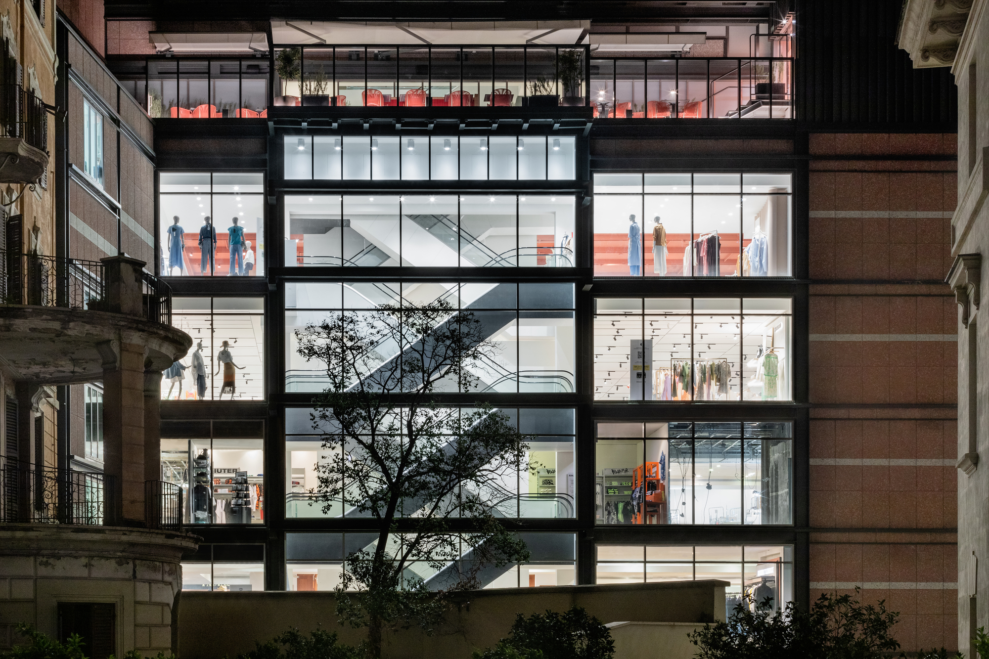 Night view through full-height glazed facade revealing internal escalators and shoppers across multiple illuminated floors