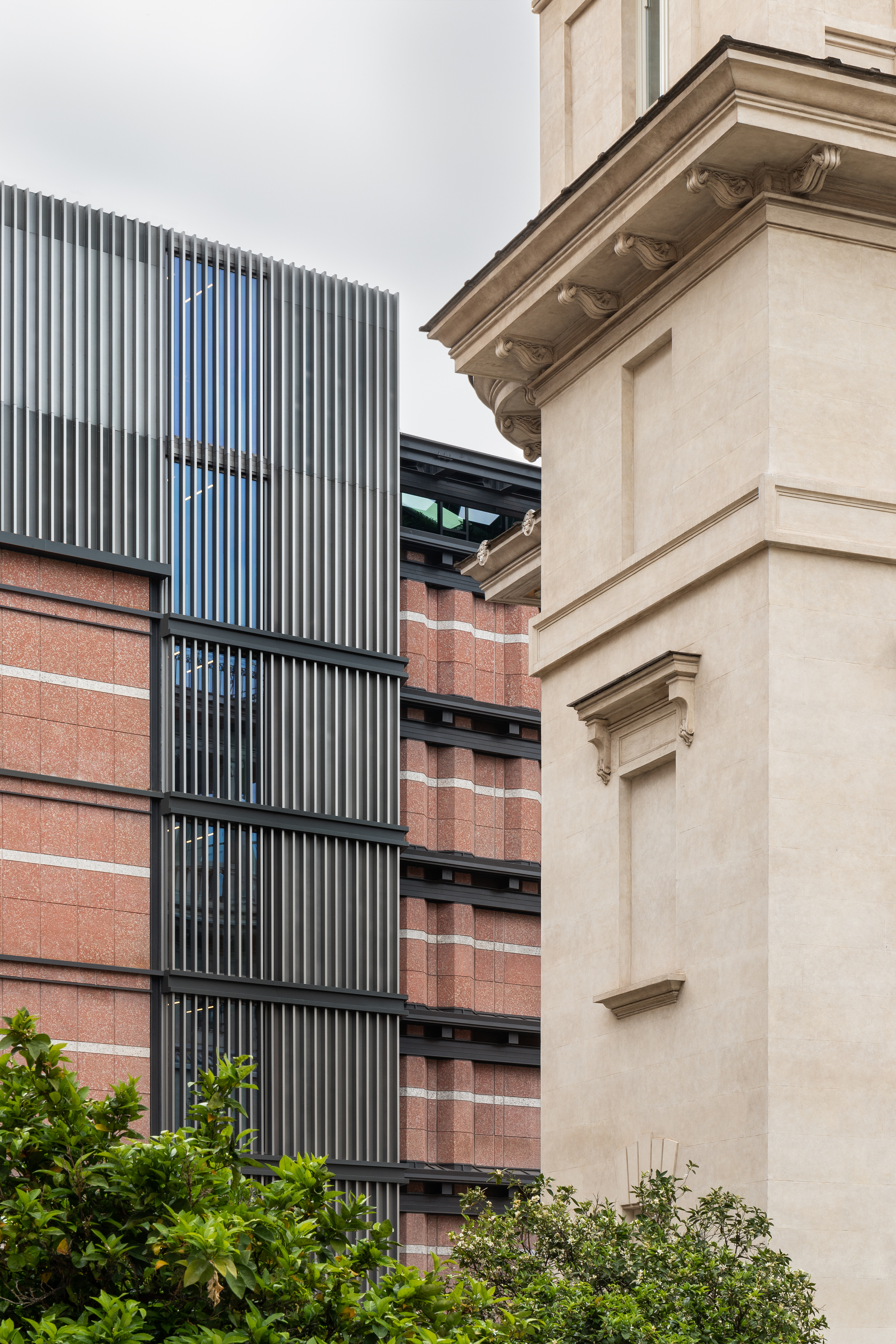 Vertical fin louvres rising beside red brick bands and a rendered heritage corner under overcast skies