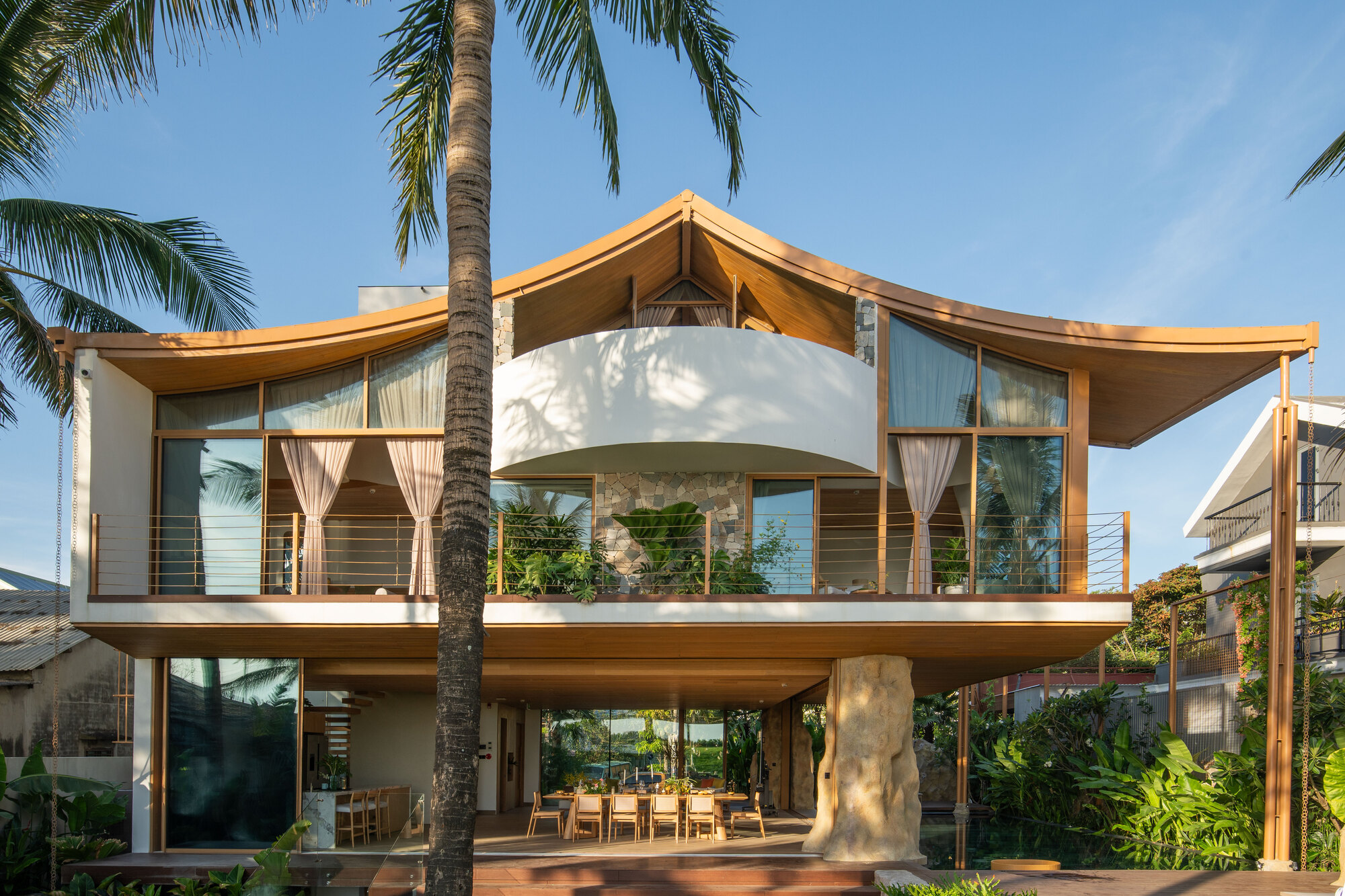 Two-story timber and white stucco facade with a curved roof sheltering a balcony and palm tree