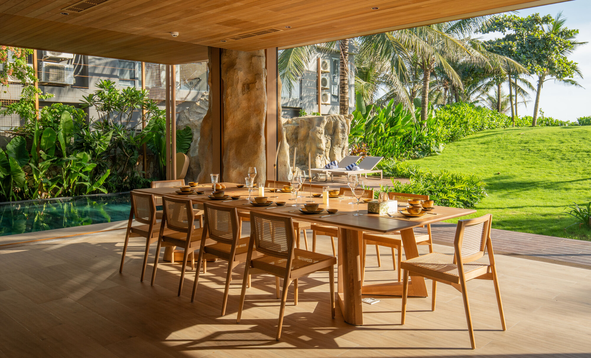 Open-air dining area beneath timber ceiling with pool and lawn visible beyond the columns