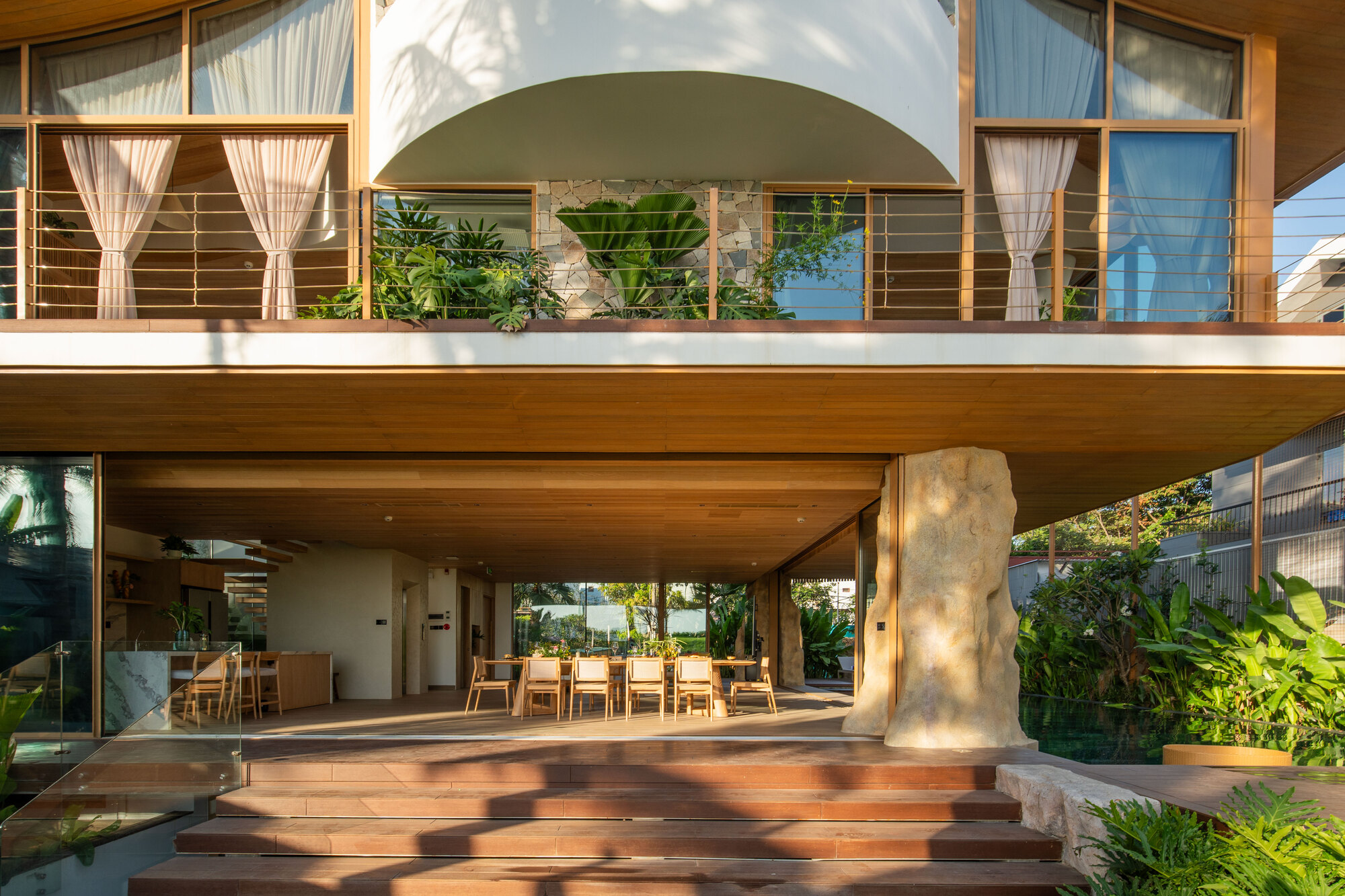 Two-level terrace with timber deck steps and cylindrical stone column beneath a deep overhang in afternoon light